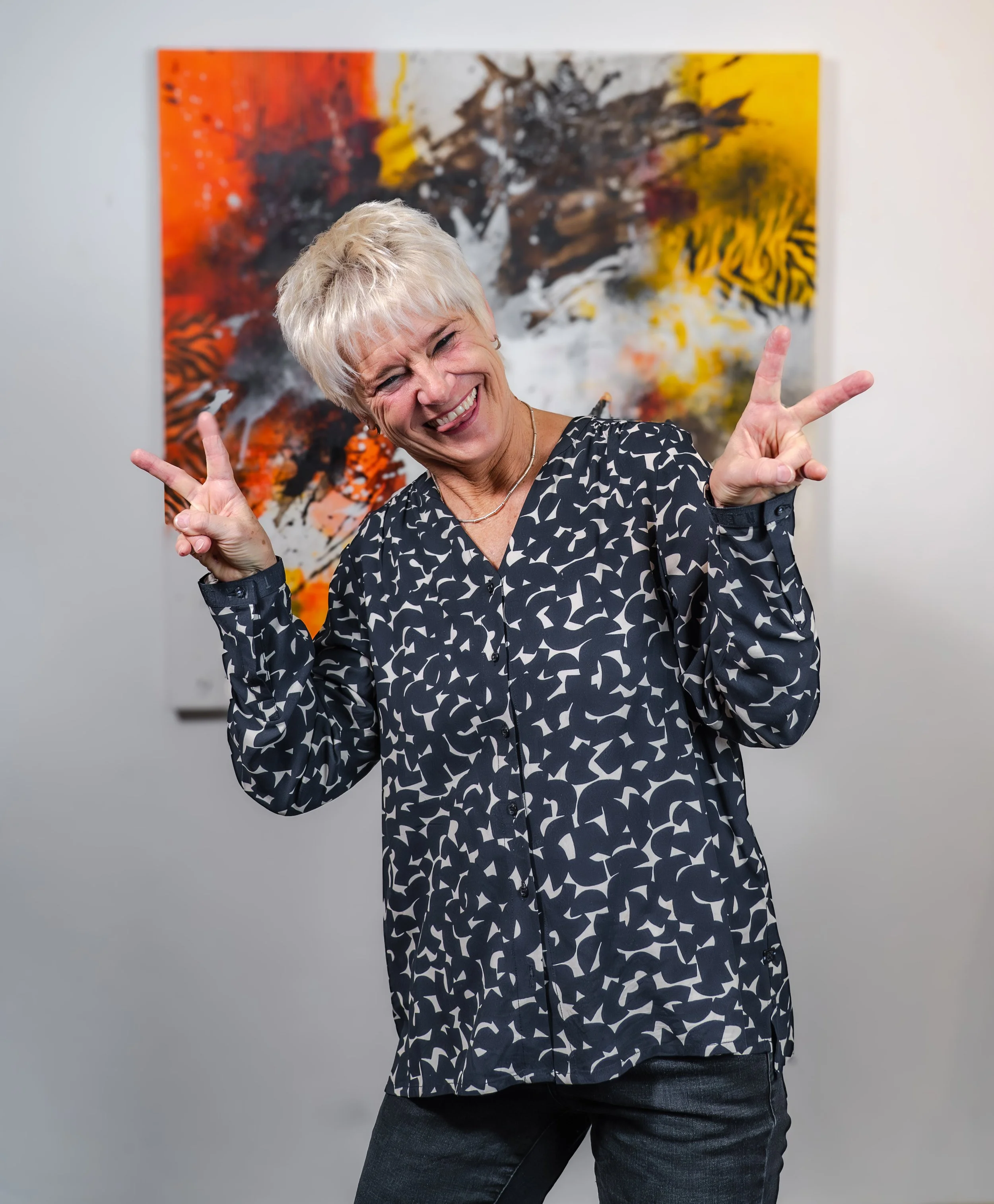 Lächelnde Frau in schwarzem und weißem Hemd macht peace-Zeichen mit beiden Händen, im Hintergrund ein abstraktes Kunstwerk mit Rot, Schwarz, Weiß und Gelb.