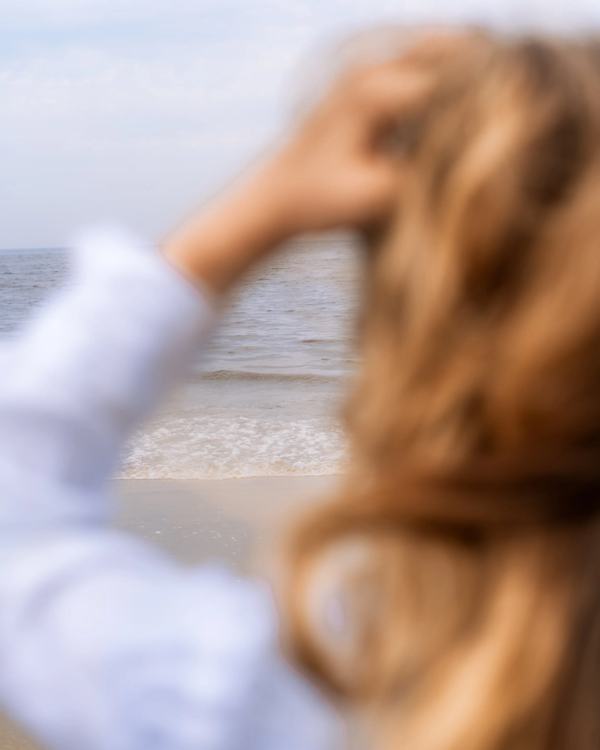 Das Bild zeigt eine Person mit langem, braunem Haar, die am Strand steht und in die Kamera schaut, während die Wellen des Meeres im Hintergrund zu sehen sind.