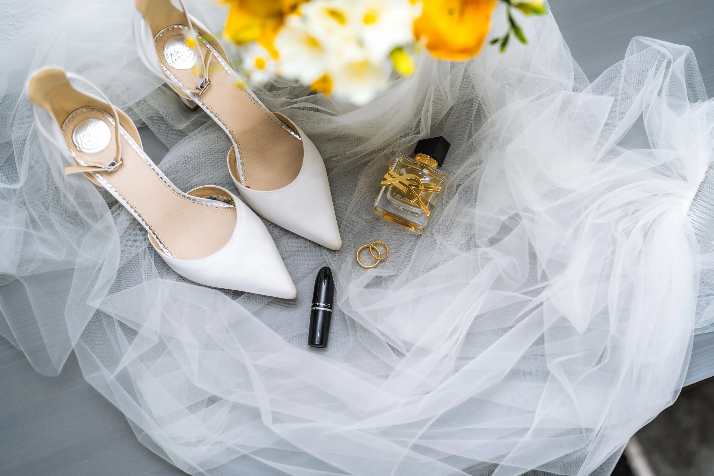 Ein Paar weiße High Heels, Parfümflasche, Lippenstift, Ohrringe, Ringe, Make-up-Stift, Blume, Tüllstoff auf einem Tisch.