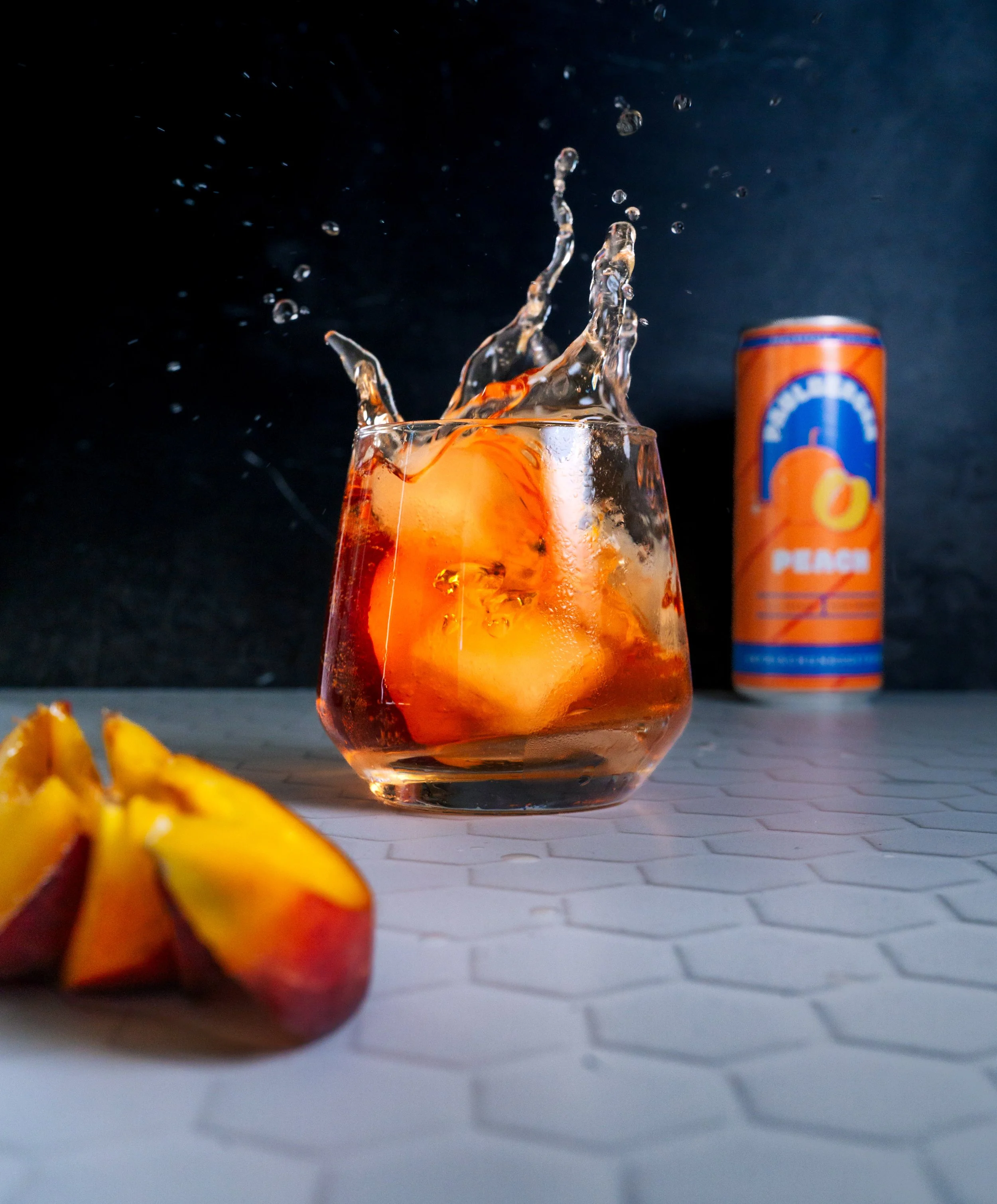 Ein Glas mit Orangenlimonade und Eiswürfeln, daneben eine Dose mit Orangenlimonade, auf einer weißen, hexagonalen Fliese. Das Wasser spritzt aus dem Glas beim Eingießen der Limonade.