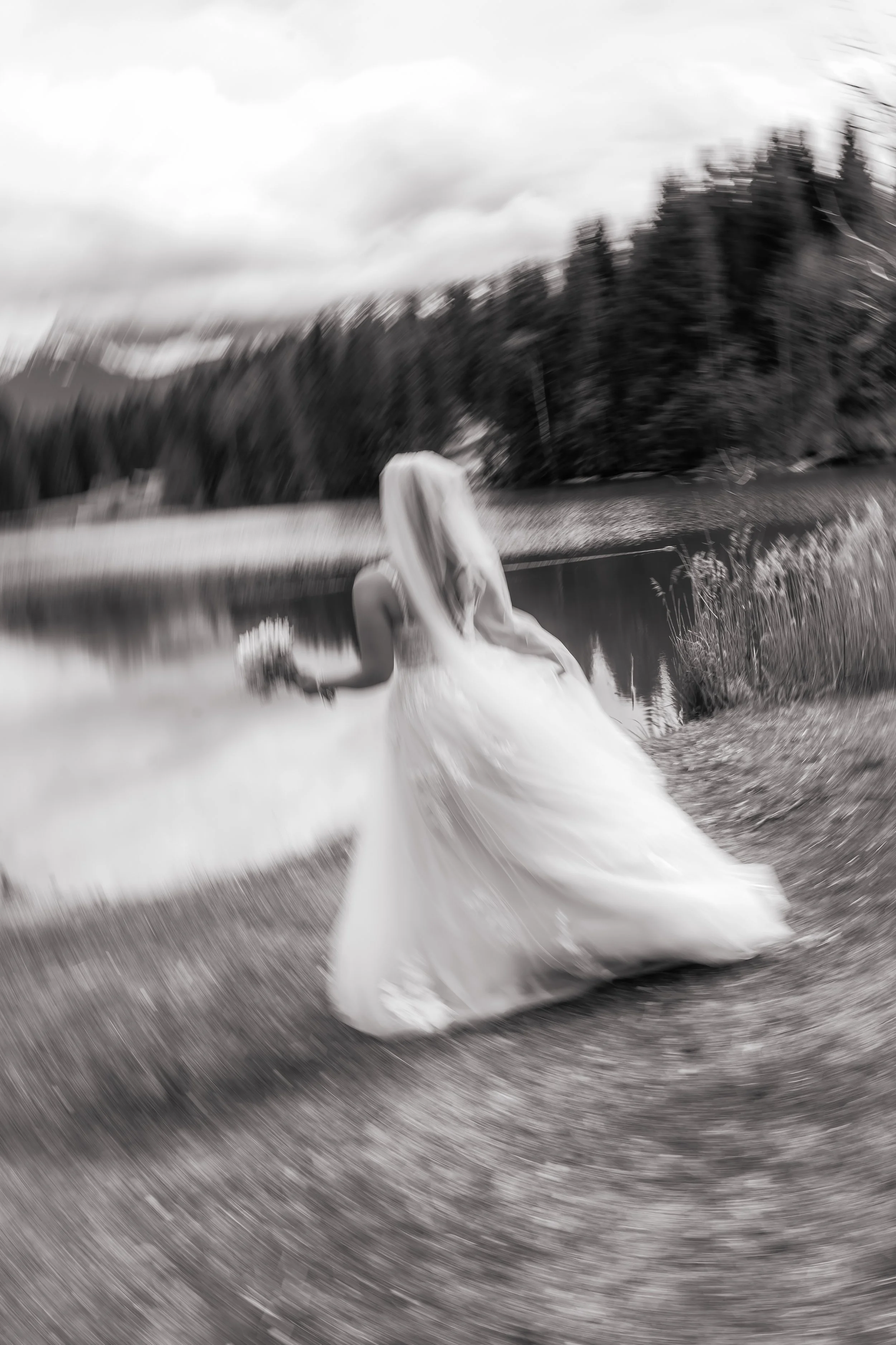 Eine Braut in einem weißen Brautkleid, die an einem Fluss oder See steht, mit einer natürlichen Landschaft im Hintergrund. Das Bild ist unscharf und in Schwarz-Weiß gehalten.