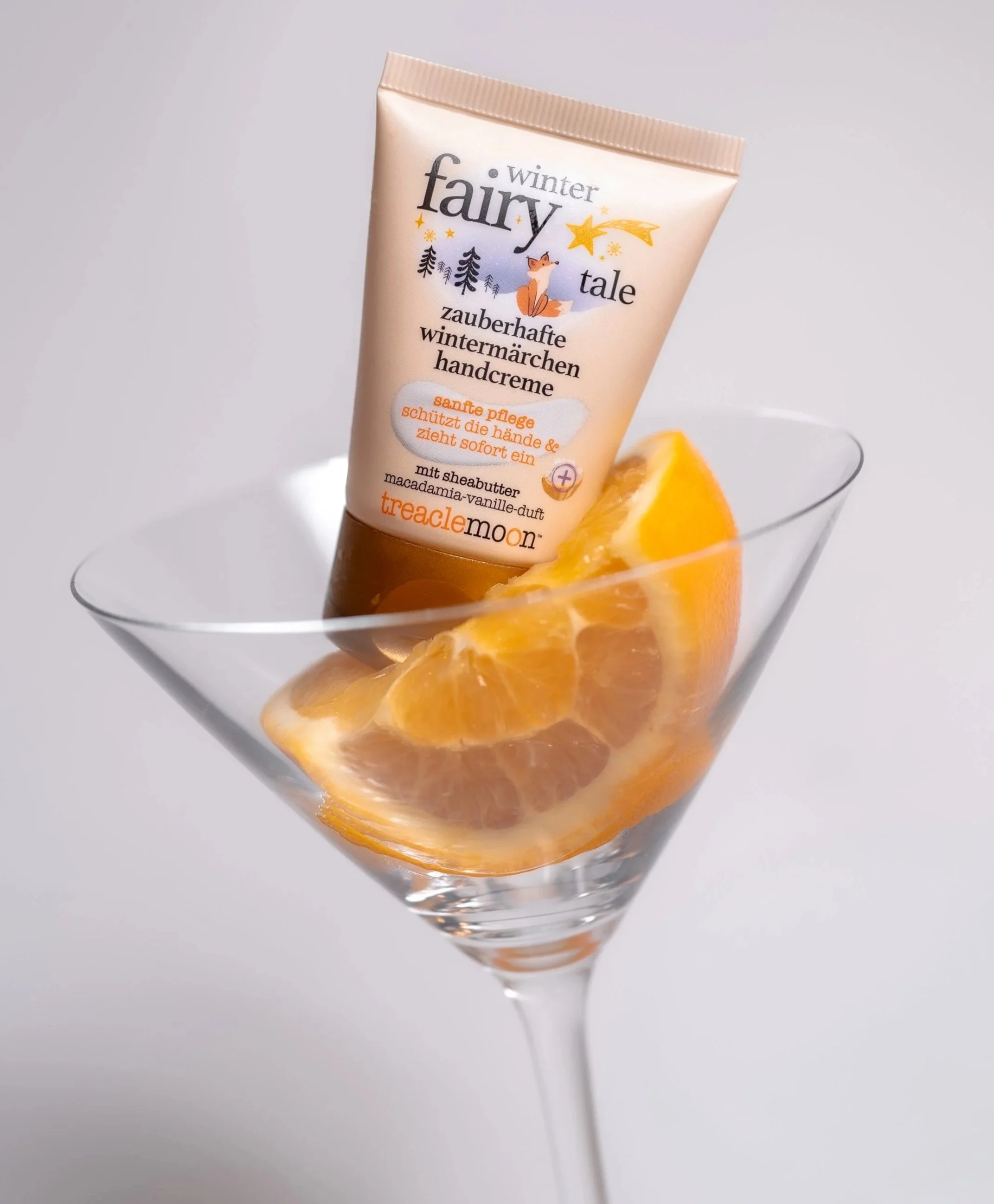 Eine Tube Handcreme steht in einem Weinglas mit orangefarbenen Orangenstücken. Die Tube trägt den Namen "Winter Fairy Tale" und hat eine winterliche Illustration mit Tannenbäumen, Sternen, einem Fuchs und einem Zauberstern.