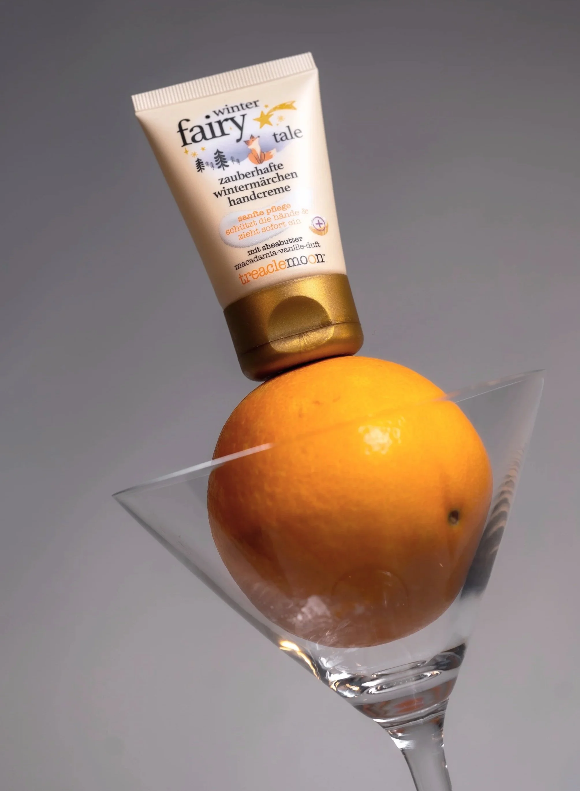 Ein Cocktailglas mit einer großen Orangel und einer Handcreme-Tube, die auf dem Apfel steht. Der Hintergrund ist grau.