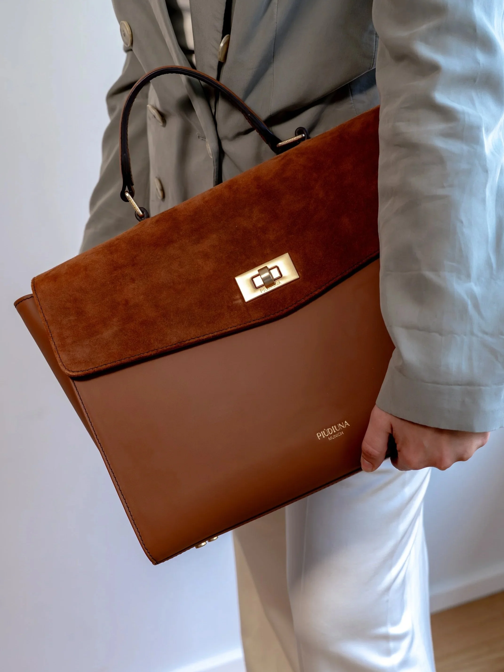 Person trägt eine braune Ledertasche mit goldener Klemme, während sie ein hellblaues Hemd und weiße Hosen trägt.