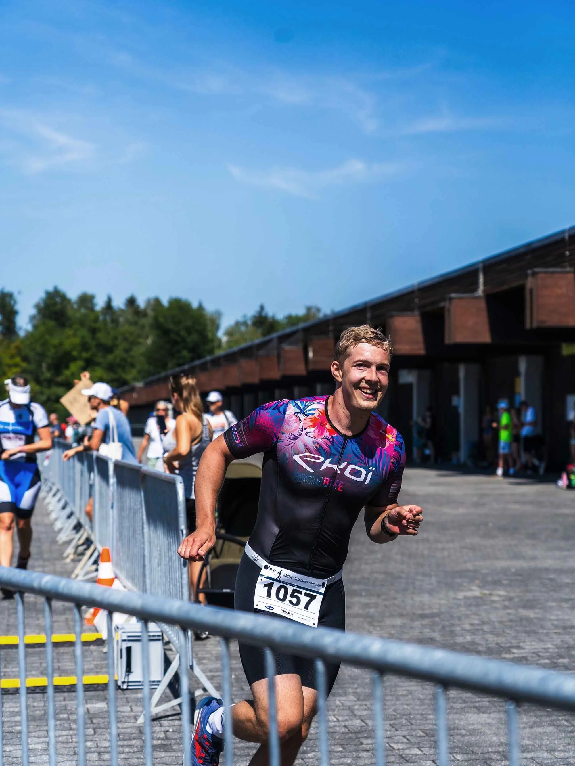 Mann beim Marathonlauf, trägt ein buntes Laufshirt, läuft auf einer Straße mit Barrieren und Zuschauern im Hintergrund an einem sonnigen Tag.