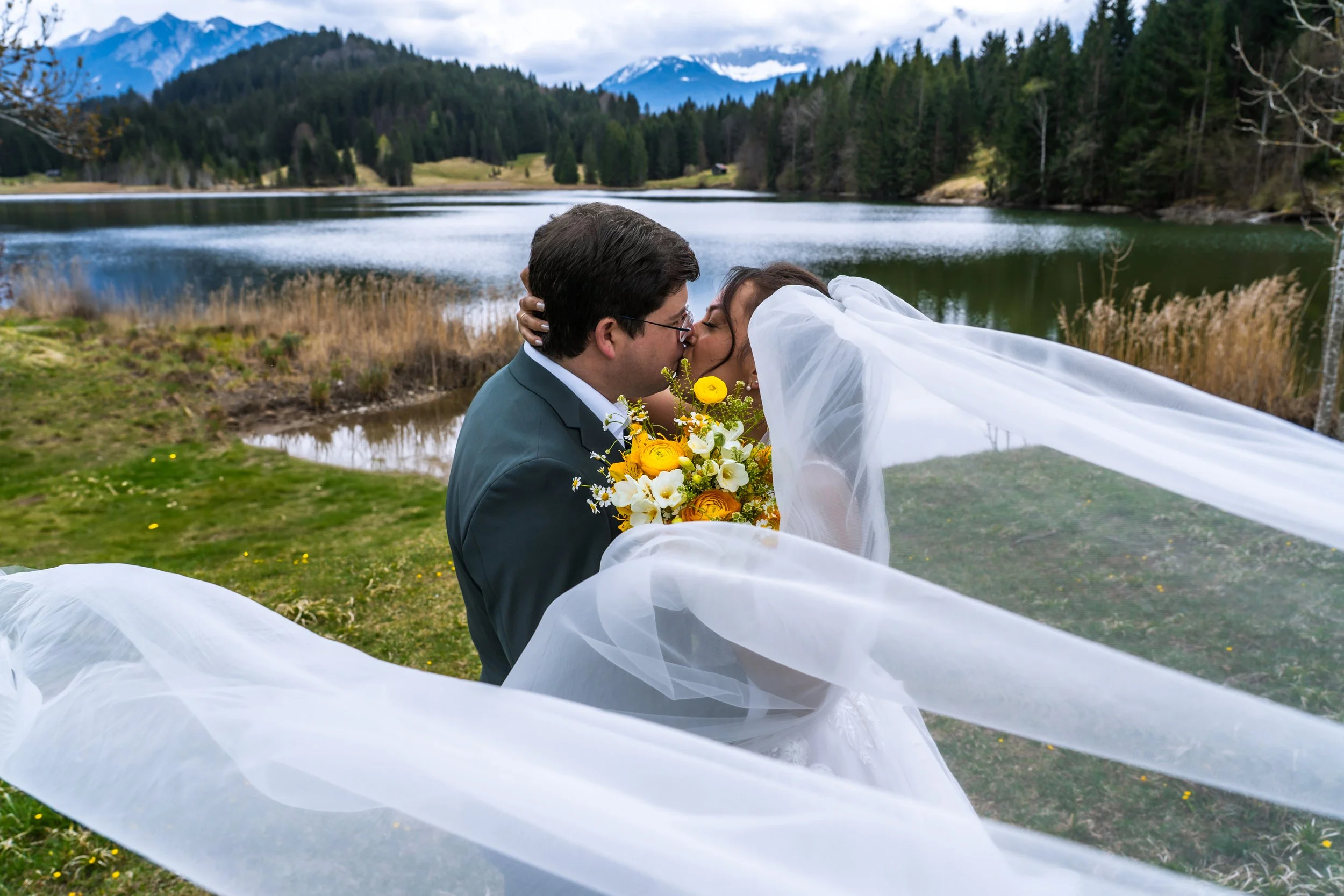 Ein Brautpaar bei ihrer Hochzeit küsst sich am Ufer eines Sees mit Bergen und Wäldern im Hintergrund.