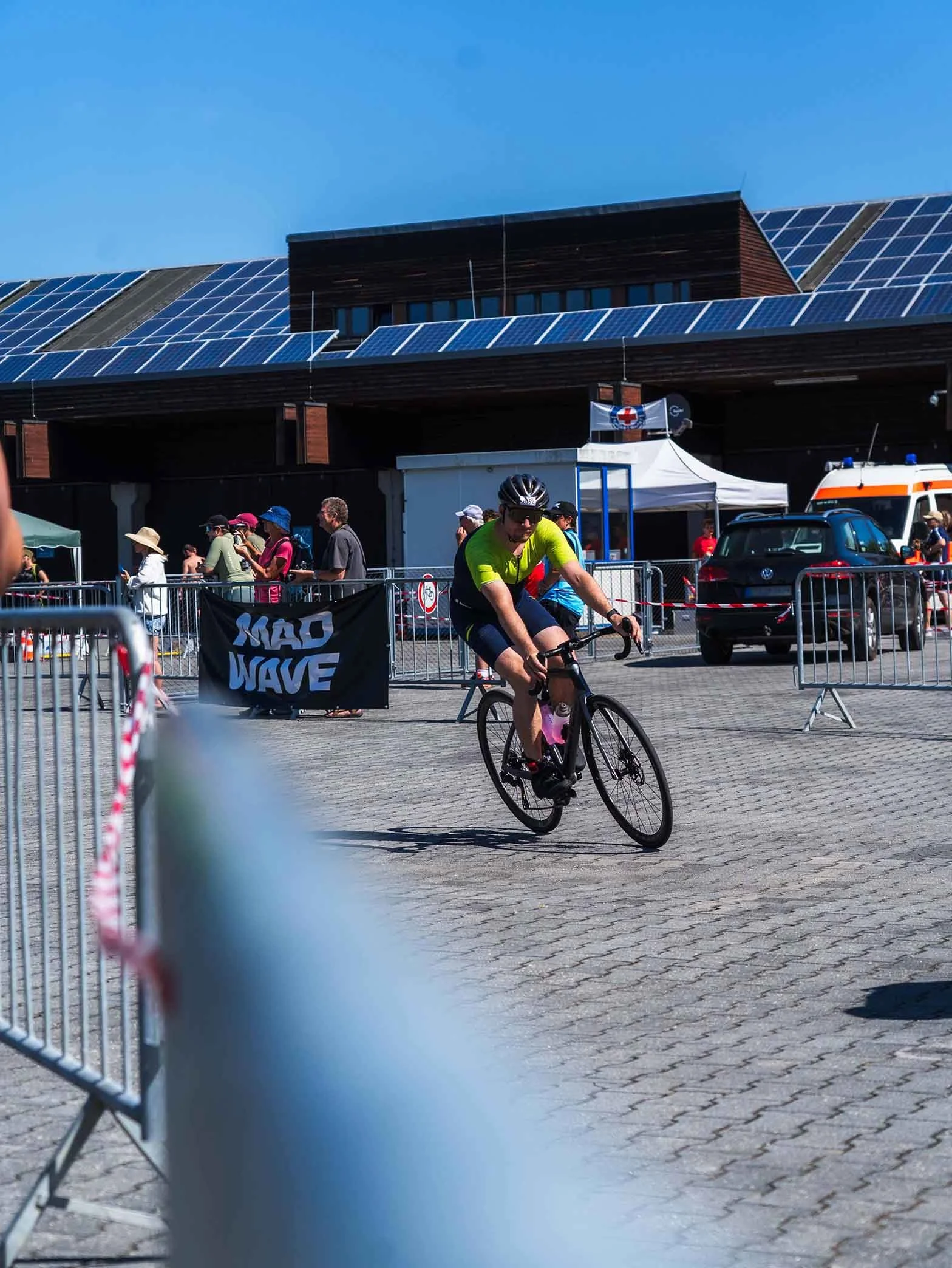 Ein Fahrradfahrer in sportlicher Kleidung fährt in einem Rennen auf einer gepflasterten Straße, im Hintergrund sind Menschen, Zelte, ein Krankenwagen, und ein Gebäude mit Solarmodulen auf dem Dach bei sonnigem Himmel.