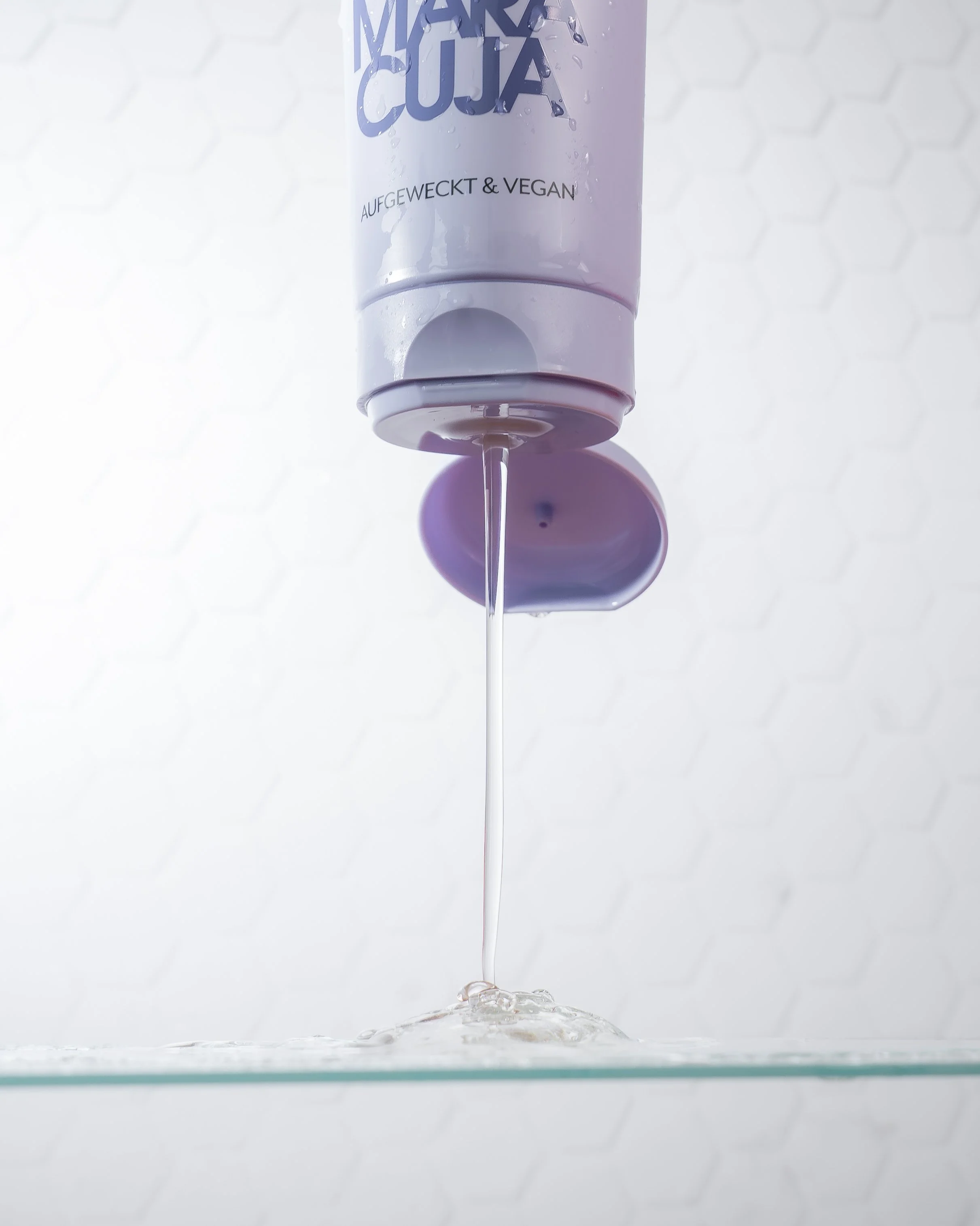 Ein Duschgel in einer Dusche, aus der Flüssigkeit auf eine Glasablage in der Dusche tropft, vor einem weißen, hexagonalen Hintergrund.