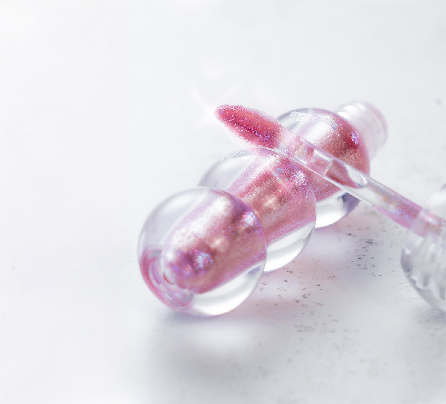 Ein naßes Lippenpflegestift mit pink-glitzerndem Lipgloss-Produkt in einer Glasampulle auf weißem Hintergrund.