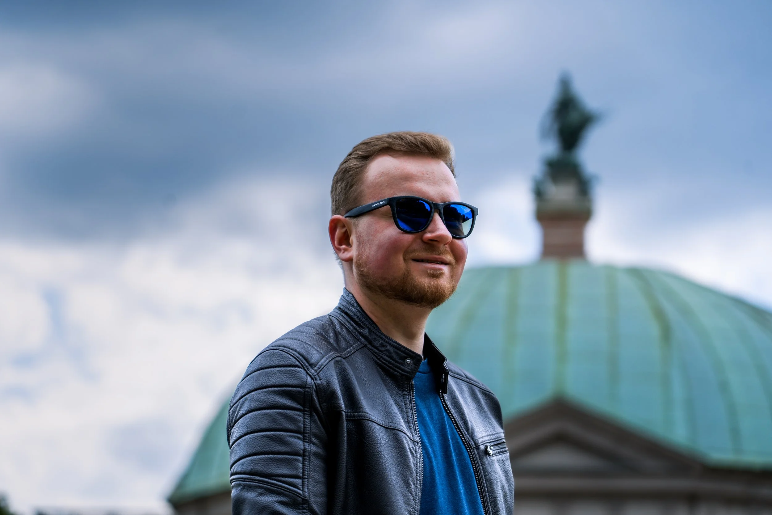 Junge Mann mit braunem Haar und Bart trägt dunkle Sonnenbrille, eine Lederjacke und ein blaues T-Shirt, steht am Odeonsplatz in München im Hofgarten und schaut lächelnd nach rechts. Der Himmel ist bewölkt.