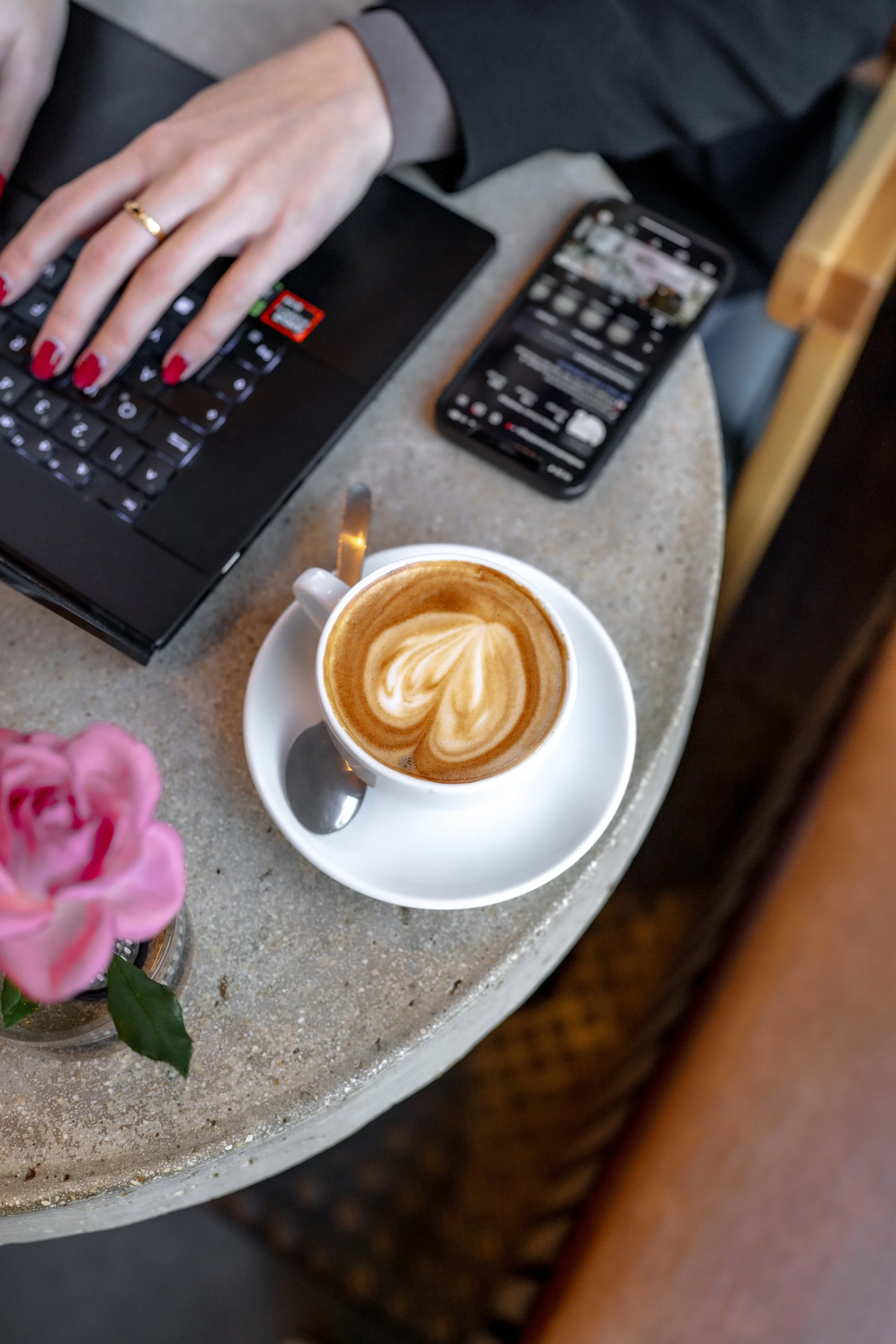 Eine Tasse Kaffee mit Milchschaum im Vordergrund, ein Laptop, ein Smartphone und eine Vase mit rosa Blumen auf einem Tisch im Hintergrund.