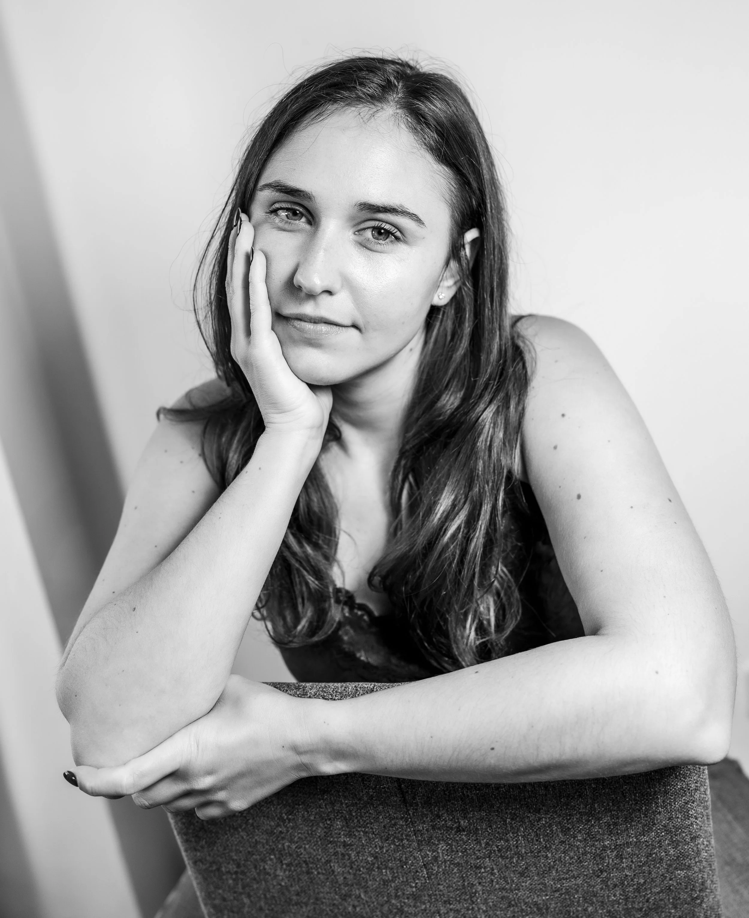 Schwarz-Weiß-Foto einer jungen Frau mit lange, lockiger Haare, die mit der Hand ihr Gesicht berührt, entspannt und nachdenklich aussehende Pose, sichtbar im Brustbereich, auf einem einfarbigen Hintergrund.
