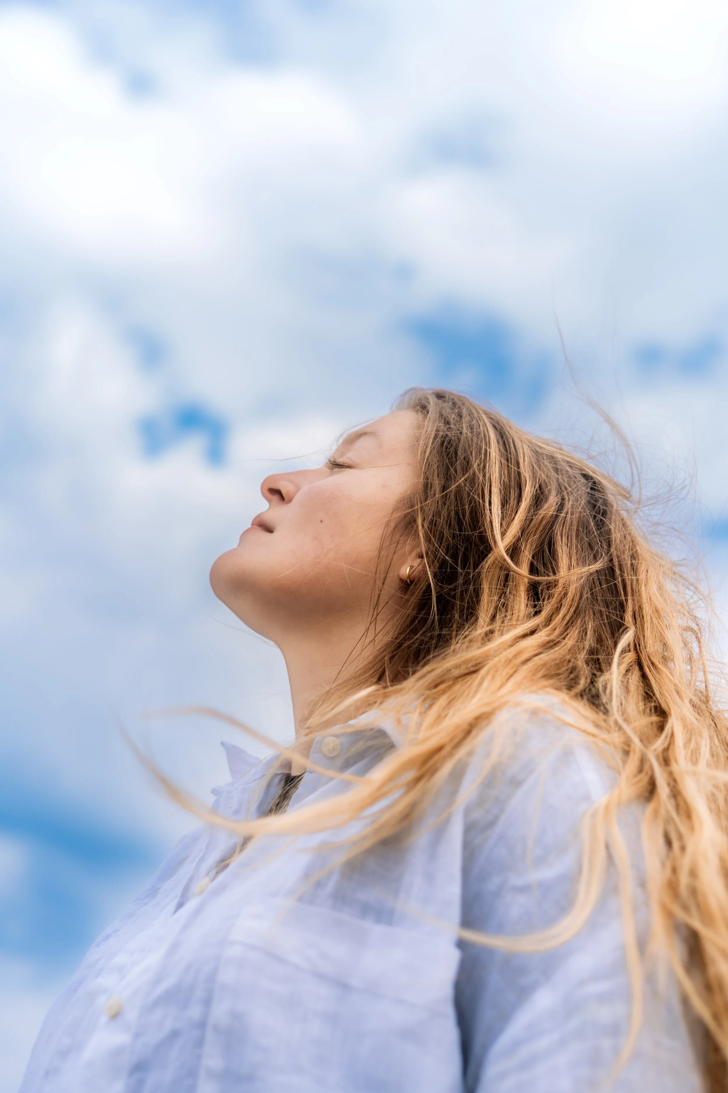 Nahaufnahme eines jungen Mädchens mit lockigem, braunem Haar, das in die Ferne schaut, bei sonnigem Himmel mit wenigen Wolken im Hintergrund.
