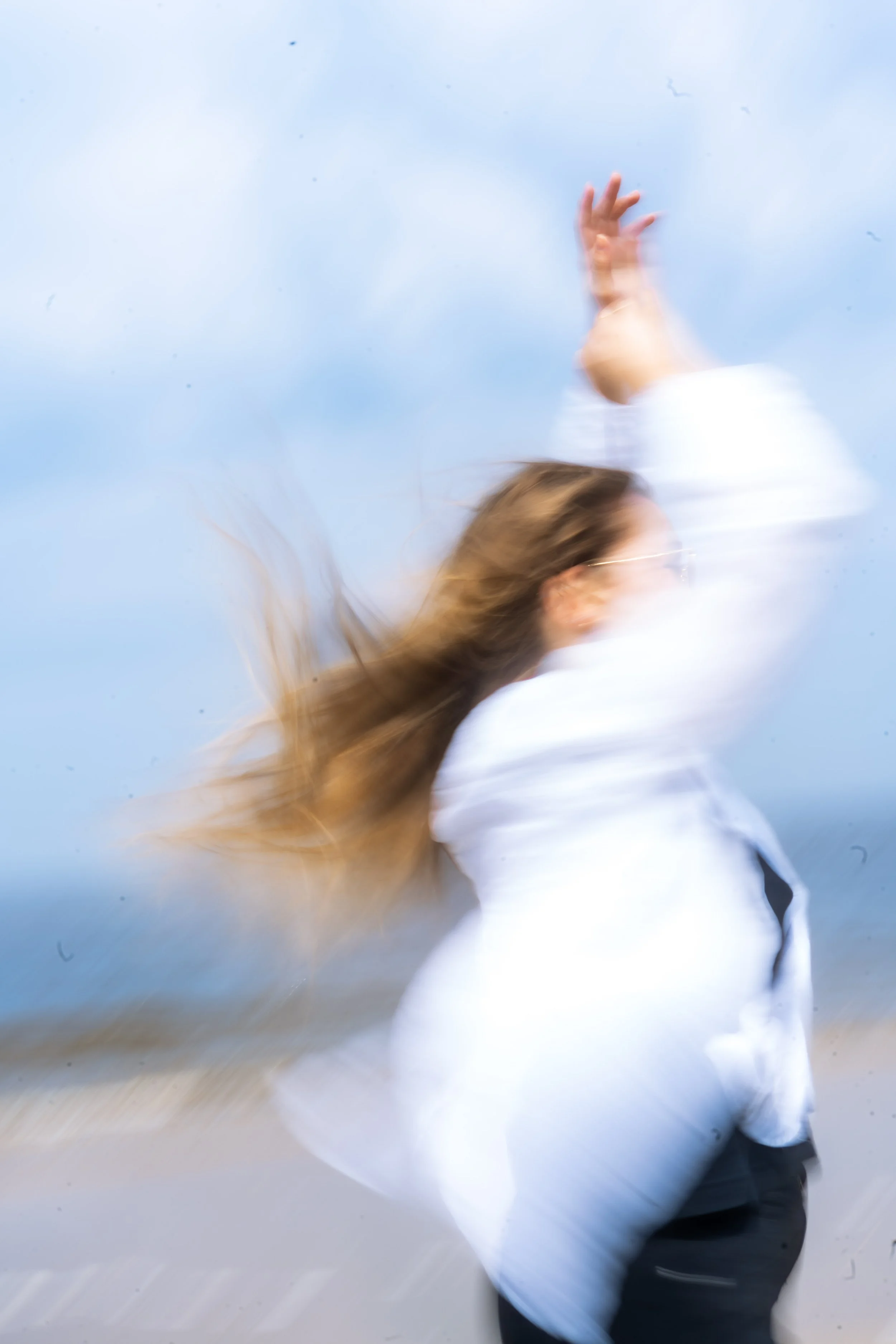 Verwackeltes Bild einer Person mit langen Haaren, die nach hinten geworfen werden, trägt eine weiße Jacke und schwarze Hose, vor einem blauen Himmel.