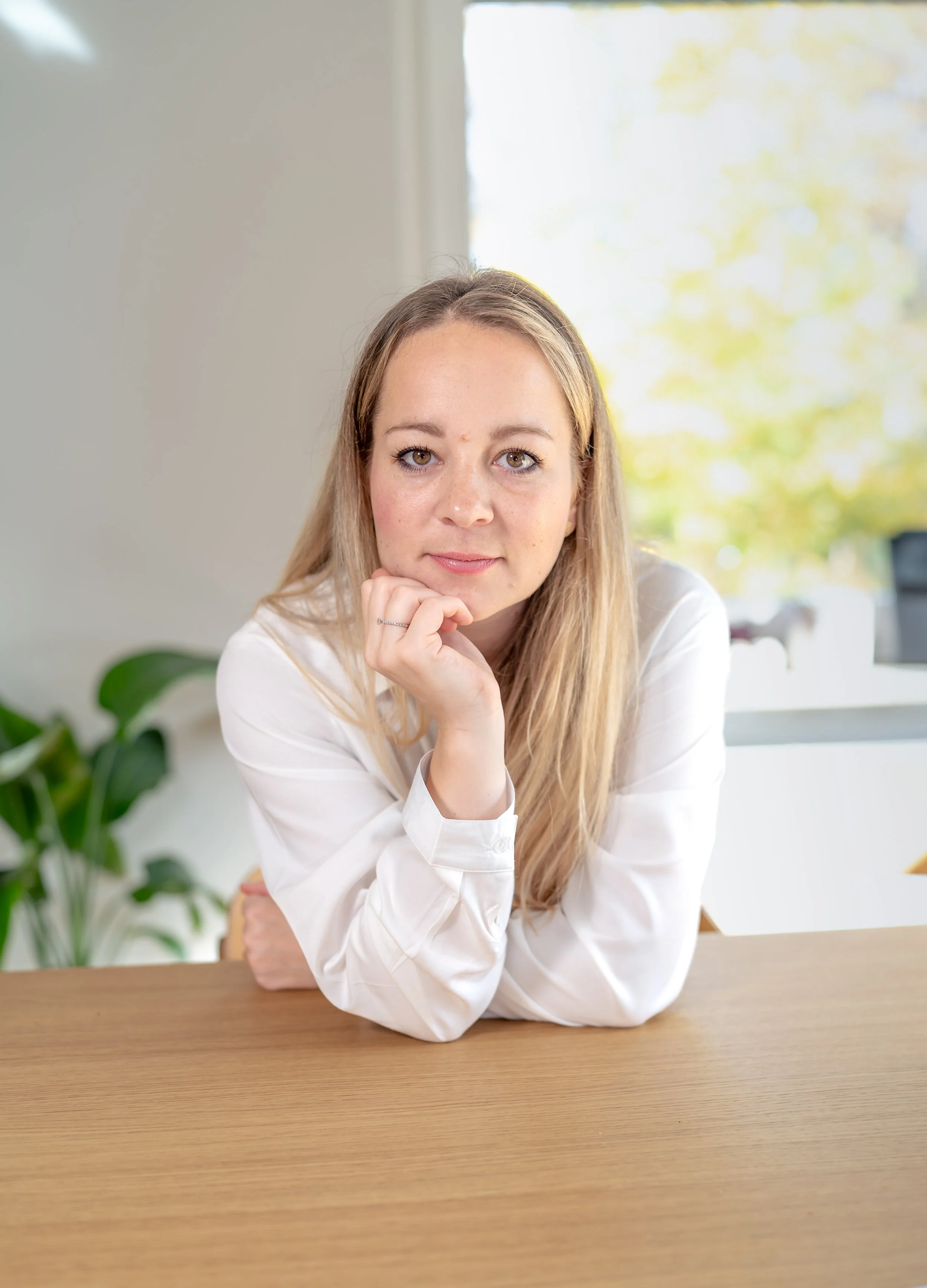 Junge Frau mit blonden Haaren und blauen Augen sitzt an einem Holztisch vor einem Fenster, legt ihre rechte Hand an ihr Kinn und schaut in die Kamera, im Hintergrund grüner Pflanzen und bunte Blätter.
