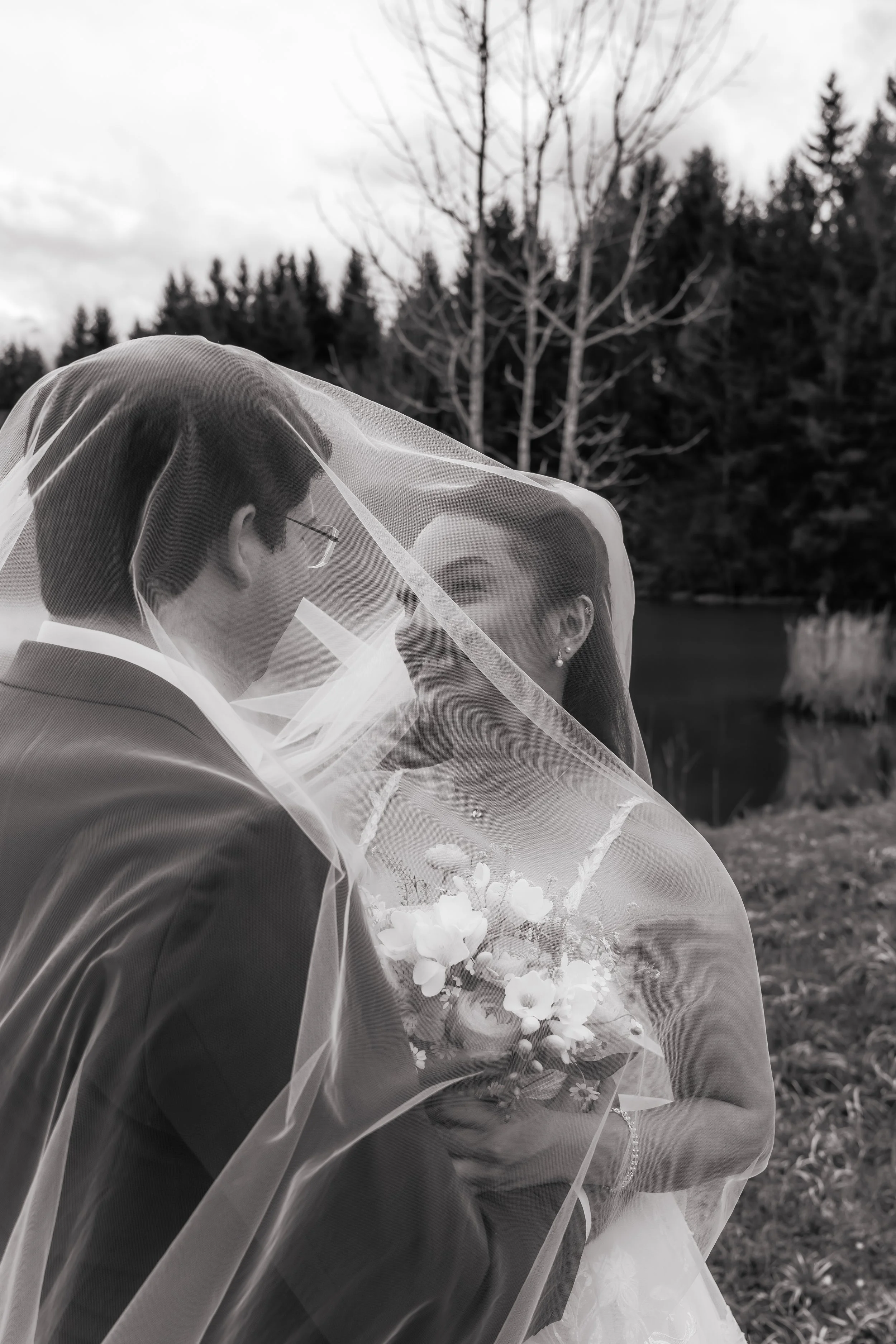 Ein Brautpaar, das sich bei einer Hochzeit im Freien küsst, während sie durch einen transparenten Schleier bedeckt sind. Im Hintergrund sind Bäume und ein See zu sehen.