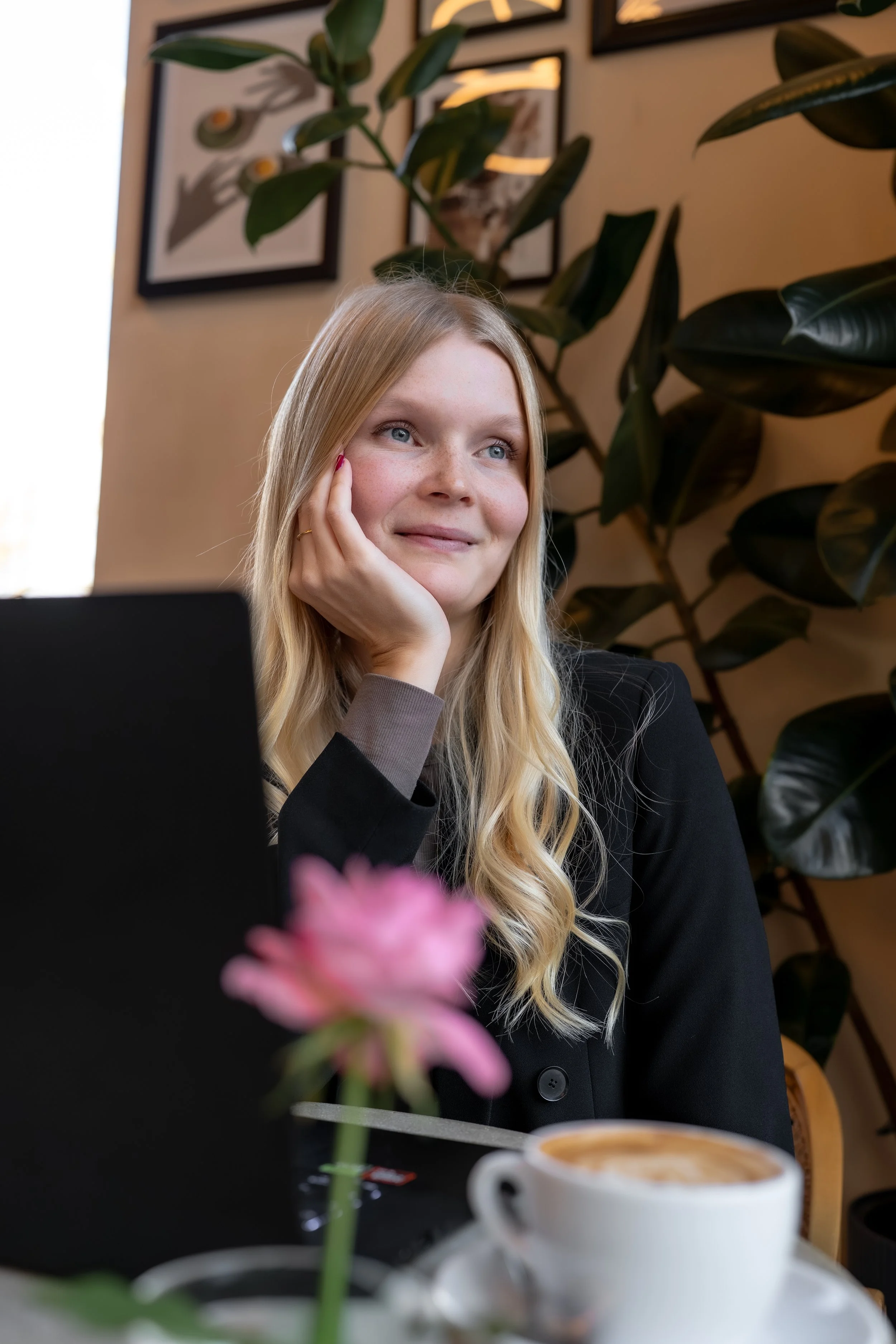 Junge blonde Frau mit blauen Augen sitzt in einem Café, schaut nach oben, während sie ihre Hand an ihr Gesicht legt. Im Vordergrund befindet sich eine Tasse Kaffee auf einem Tablett, im Hintergrund Pflanzen und Bilder.