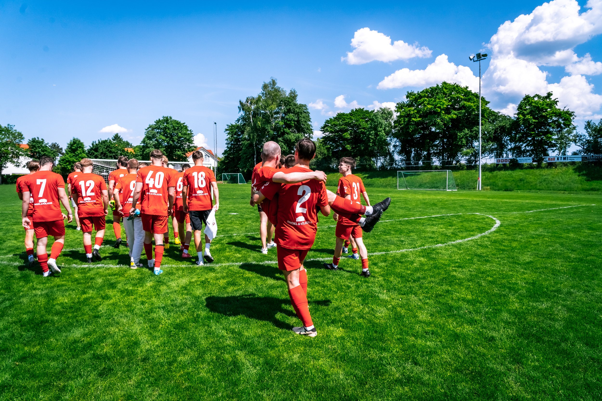 Fußballmannschaft in roten Trikots auf dem grünen Fußballfeld nach einem Spiel. Spieler umarmen sich und gehen vom Spielfeld ab. Der Himmel ist blau mit einigen Wolken, im Hintergrund sind Bäume und ein Tor zu sehen.