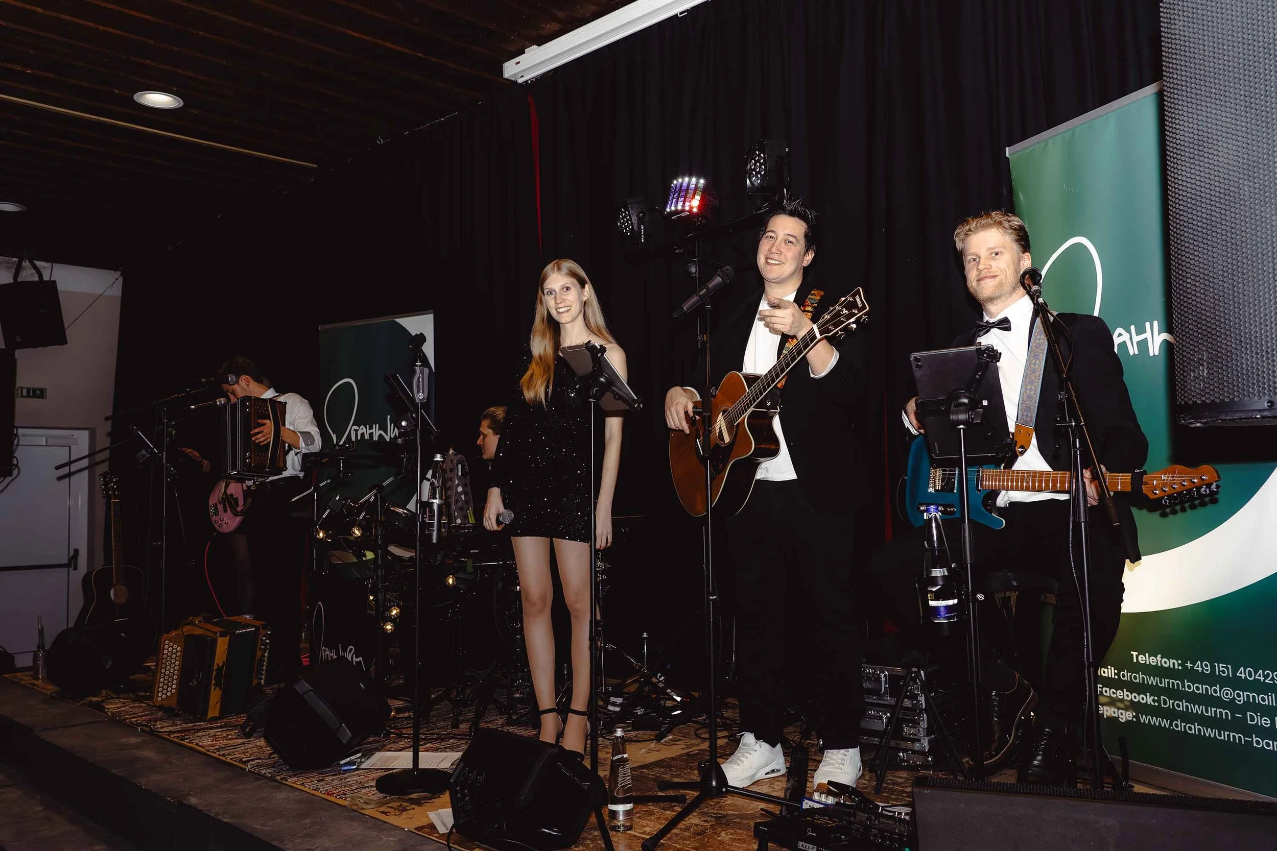 Eine Band tritt bei einer Live-Performance auf einer Bühne auf, mit einem Sänger, einer Sängerin und zwei Gitarristen, alle in formellen Outfits, mit dunklem Vorhang im Hintergrund und Bühnenbeleuchtung.