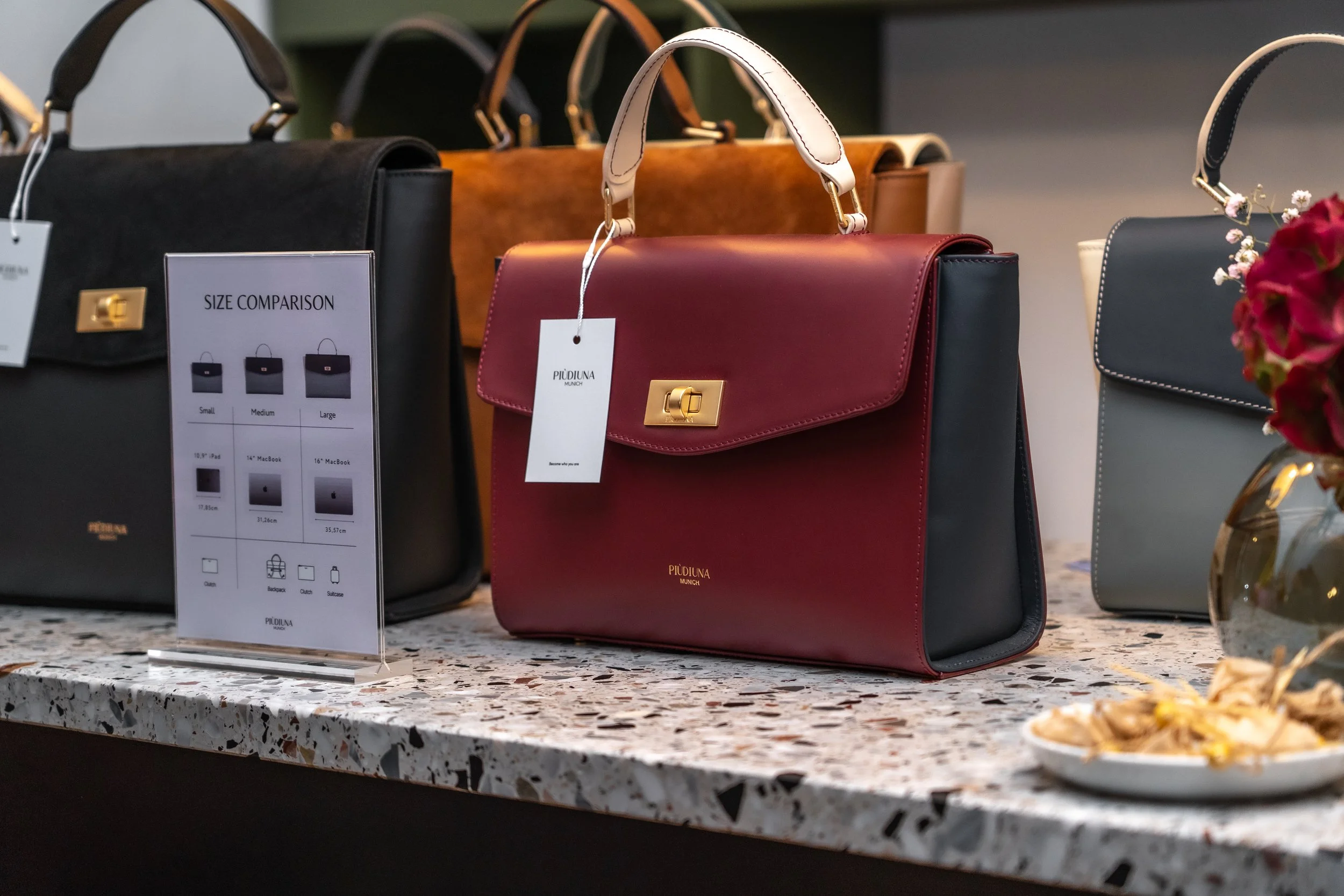 Verschiedene Handtaschen aus Leder in Schwarz, Braun, Rot und Blau auf einem Ladentisch. Ein Preisschild zeigt einen Größenvergleich für Taschen verschiedener Größen.