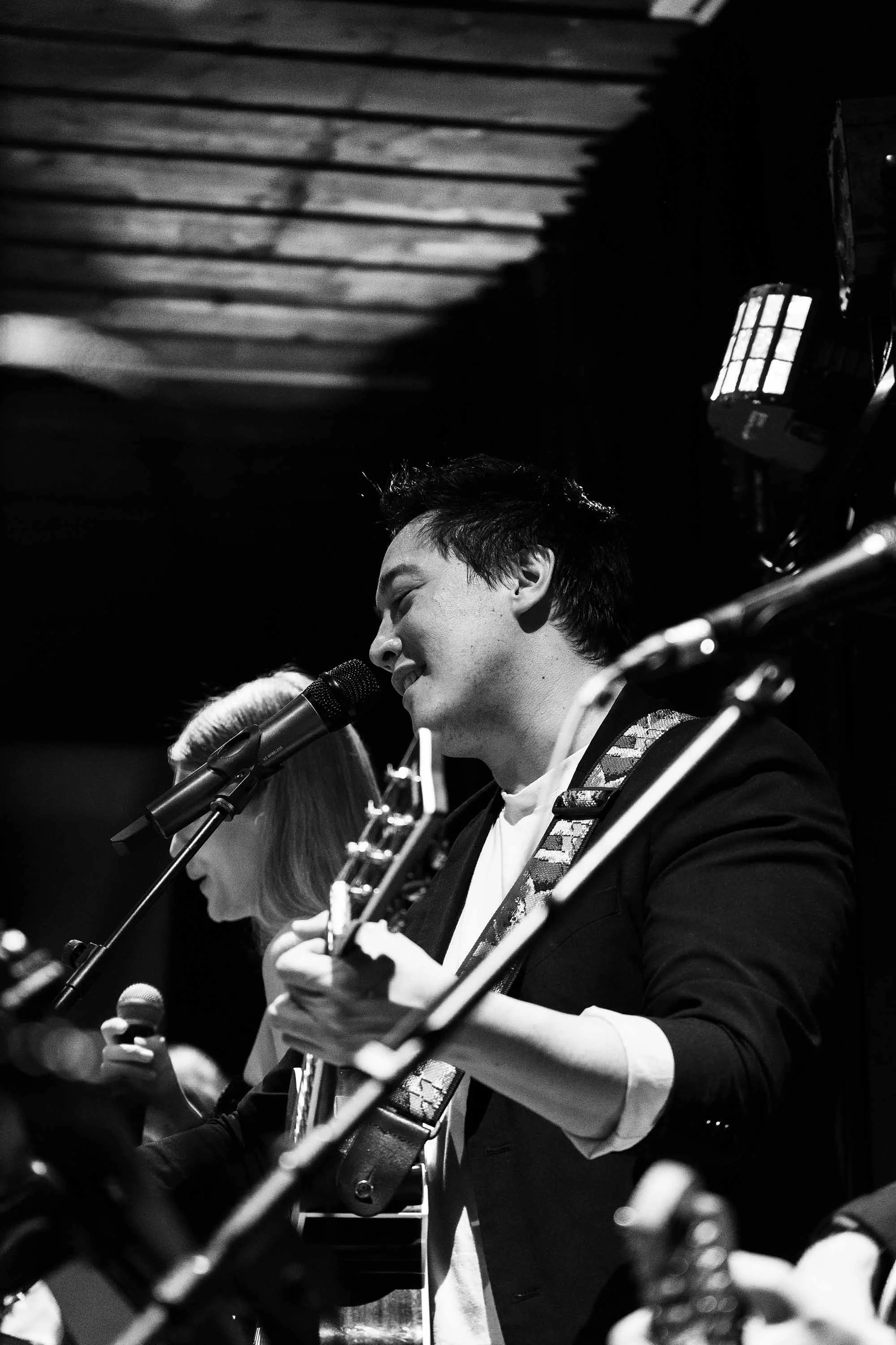 Ein junger Mann singt in einem monochromen Bild, während er Gitarre spielt und in einer Gruppe sitzt.