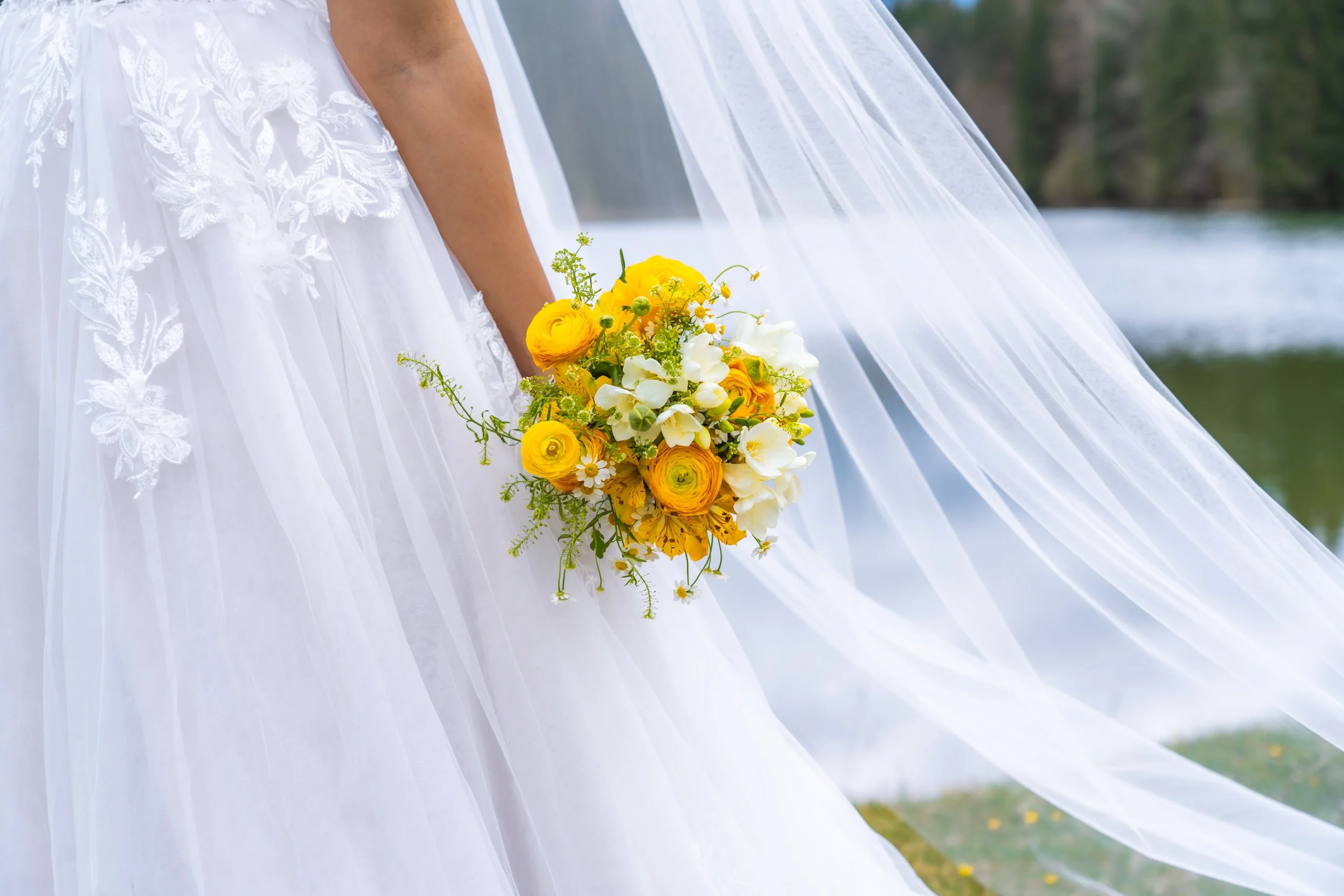 Nahaufnahme eines Brautkleids mit Spitzendetails, eine Braut hält einen gelb-weißen Blumenstrauß, im Hintergrund sind Wasser und Bäume sichtbar.