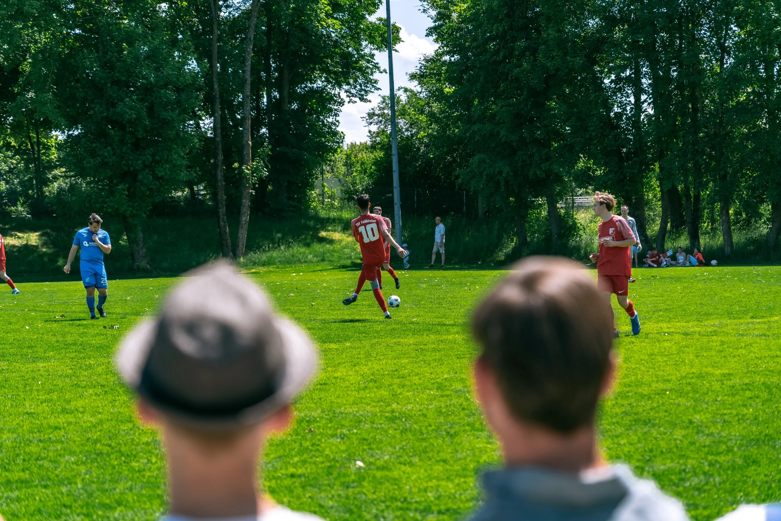Fußballteam spielen Fußball auf einer grünen Wiese, umgeben von Bäumen, Puplikum und beim Sonnenwetter.