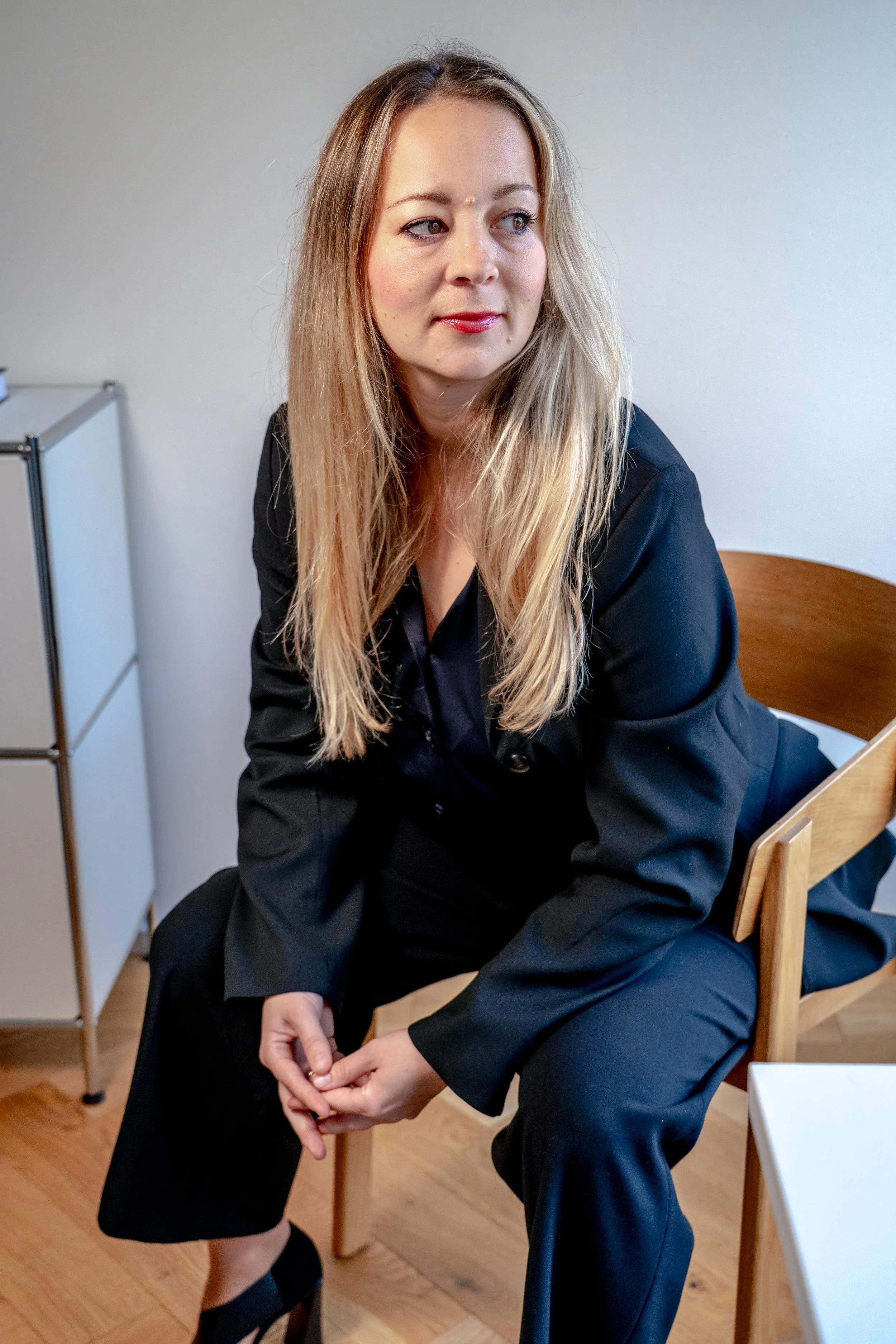 Eine Frau mit langem blonden Haar sitzt auf einem Stuhl, trägt einen schwarzen Blazer und schwarze Schuhe. Sie schaut nach rechts, mit einem leichten Lächeln. Im Hintergrund eine weiße Wand.