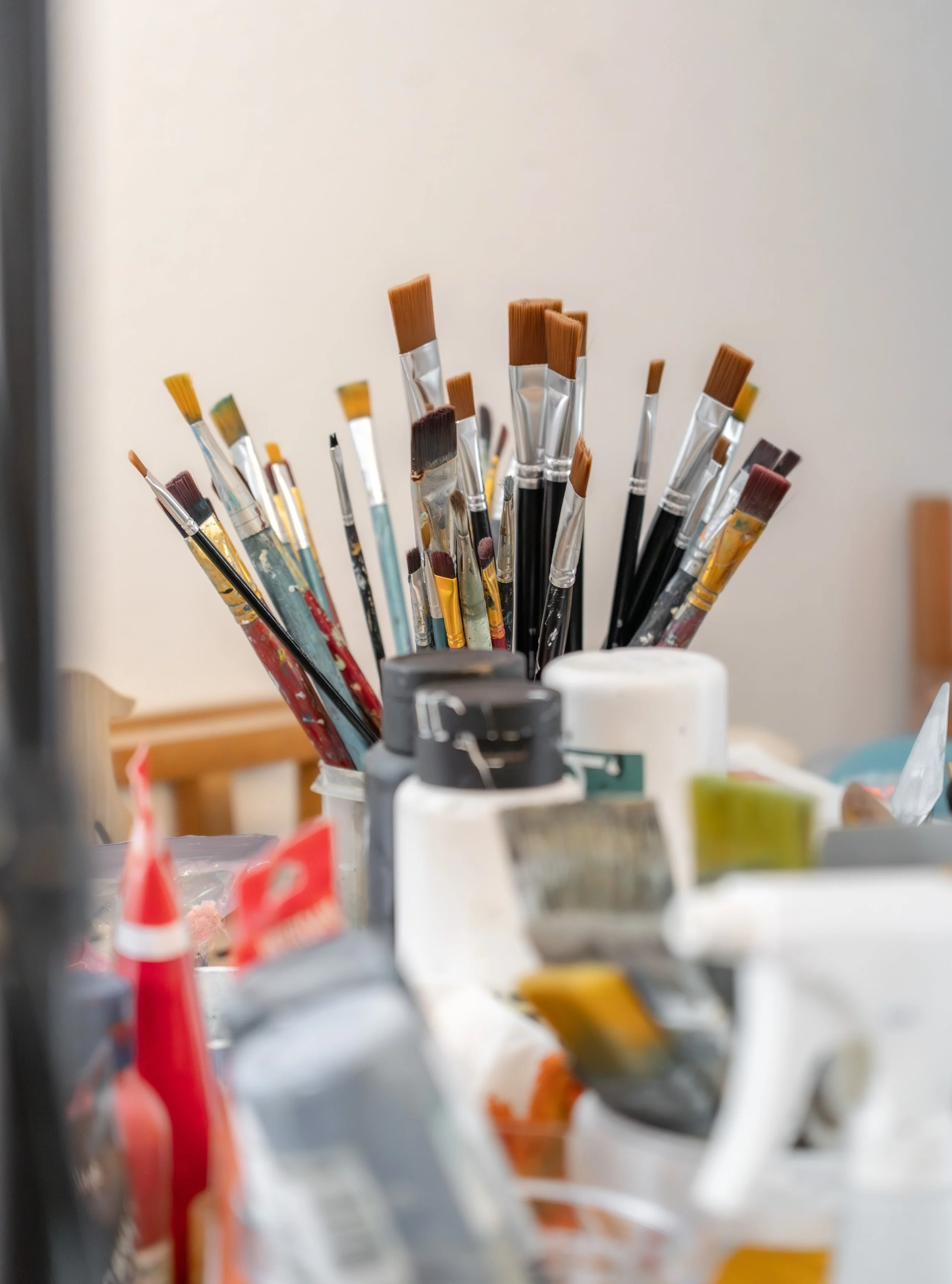 Eine Vielzahl von Pinsel unterschiedlicher Größen und Formen, einige mit Farbresten, stehen in einem Behälter auf einem Tisch, umgeben von Kunstmaterialien.