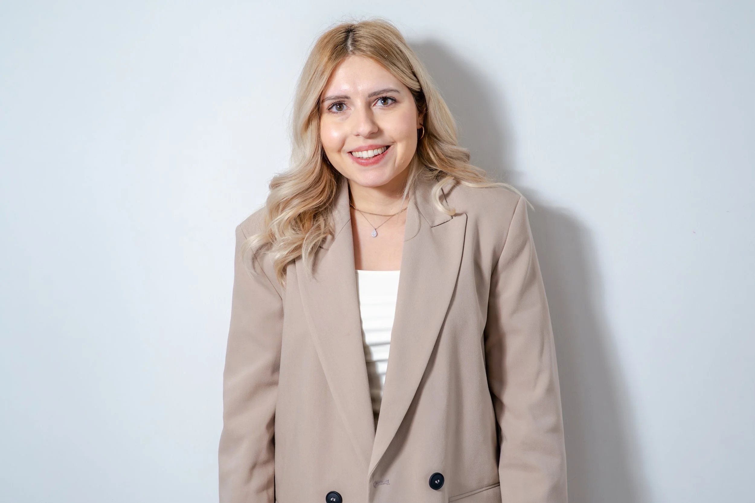 Porträt einer lachenden jungen Frau mit blonden, gewellten Haaren, in einem beige Blazer, vor hellem Hintergrund.