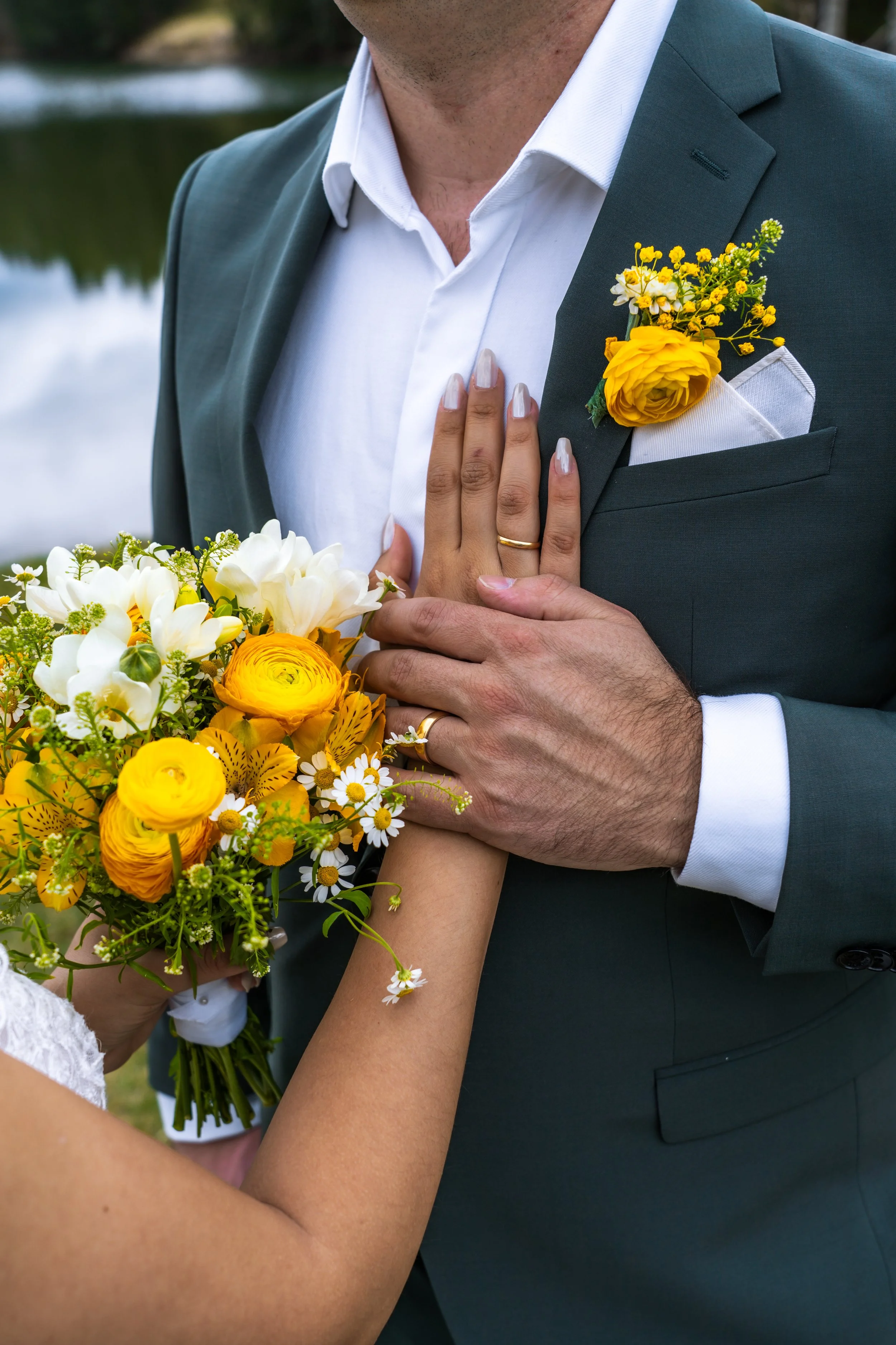 Ein Mann trägt einen dunklen Anzug mit weißem Hemd und hat einen kleinen Blumenstrauß mit gelben und weißen Blumen in der Brusttasche. Eine andere Person hält einen Blumenstrauß mit gelben, weißen und orangefarbenen Blumen.