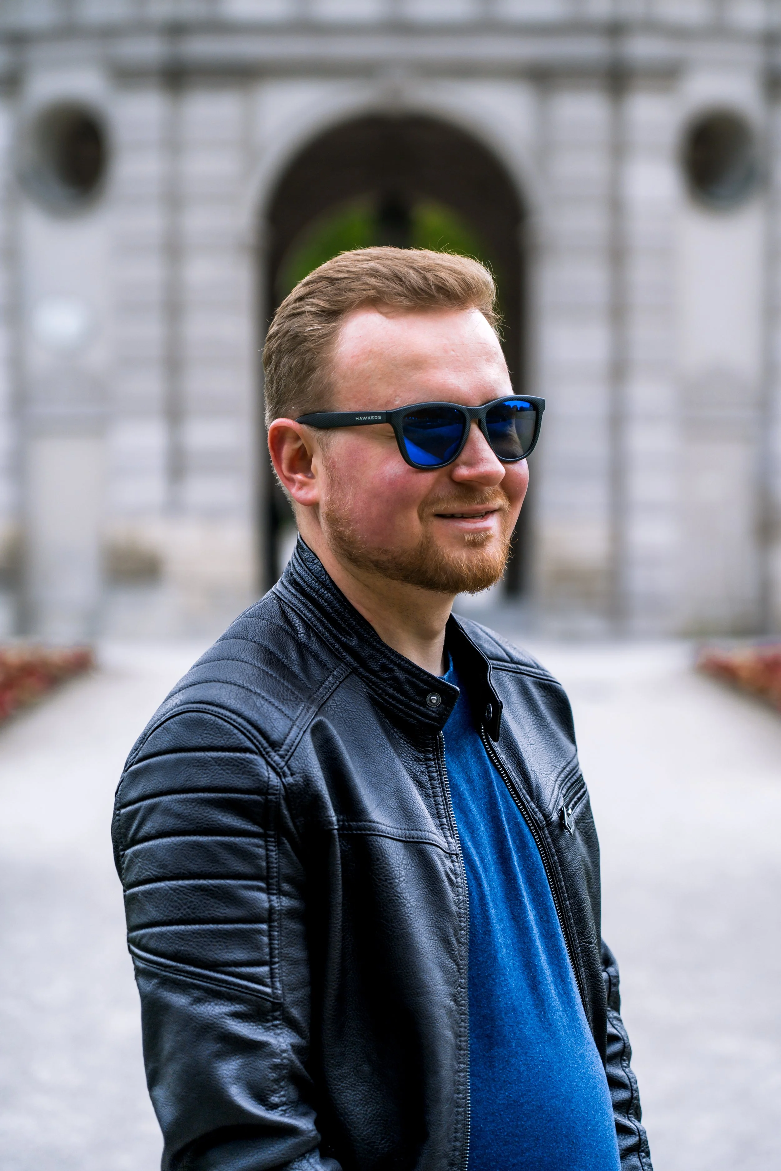 Ein Mann mit kurzen braunen Haaren, Bart und Sonnenbrille steht im Hofgarten in München vor dem Pavillion. Er trägt eine schwarze Lederjacke und einen blauen Pullover.