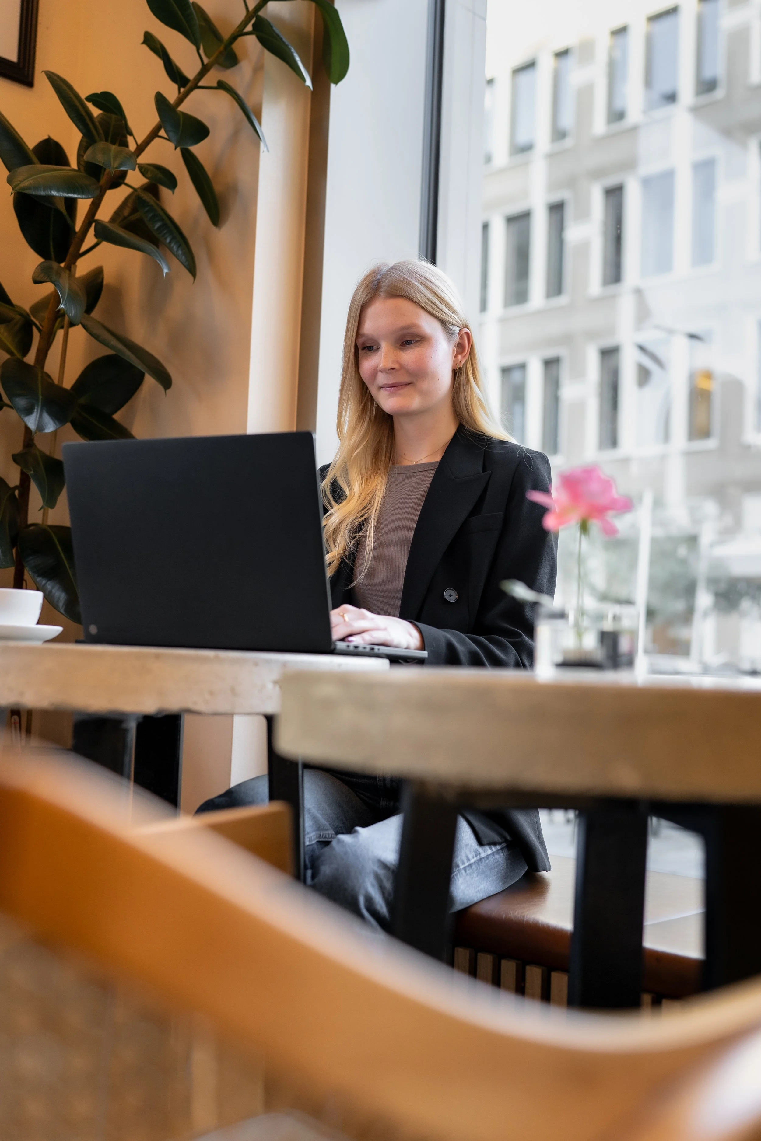 Junge Frau arbeitet an einem Laptop in einem Café mit großem Fenster, Tasse Kaffee und Blumen auf dem Tisch.