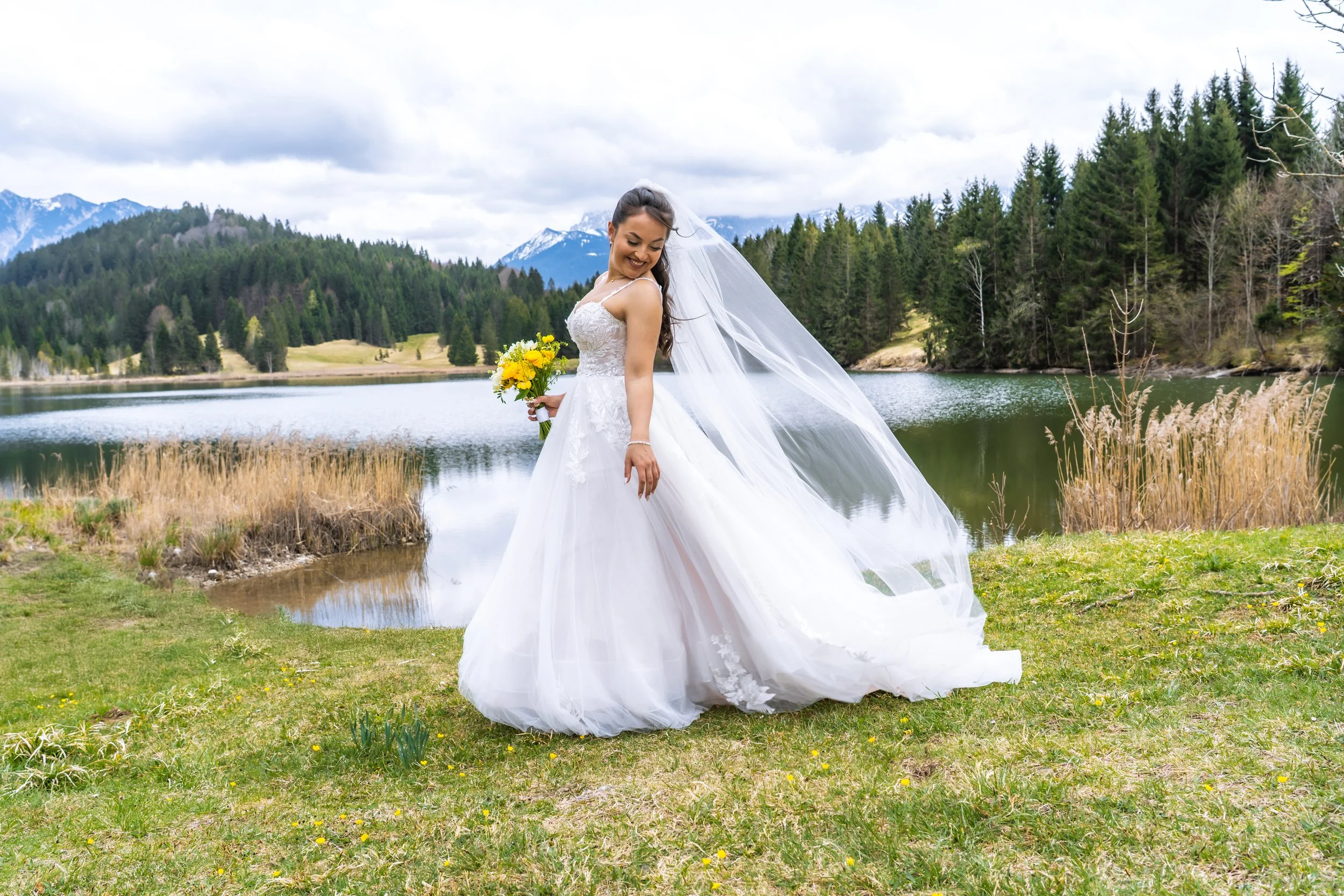 Eine lächelnde Frau in einem weißen Brautkleid mit Schleier steht an einem See in einer bergigen und bewaldeten Landschaft, hält einen gelben Blumenstrauß.