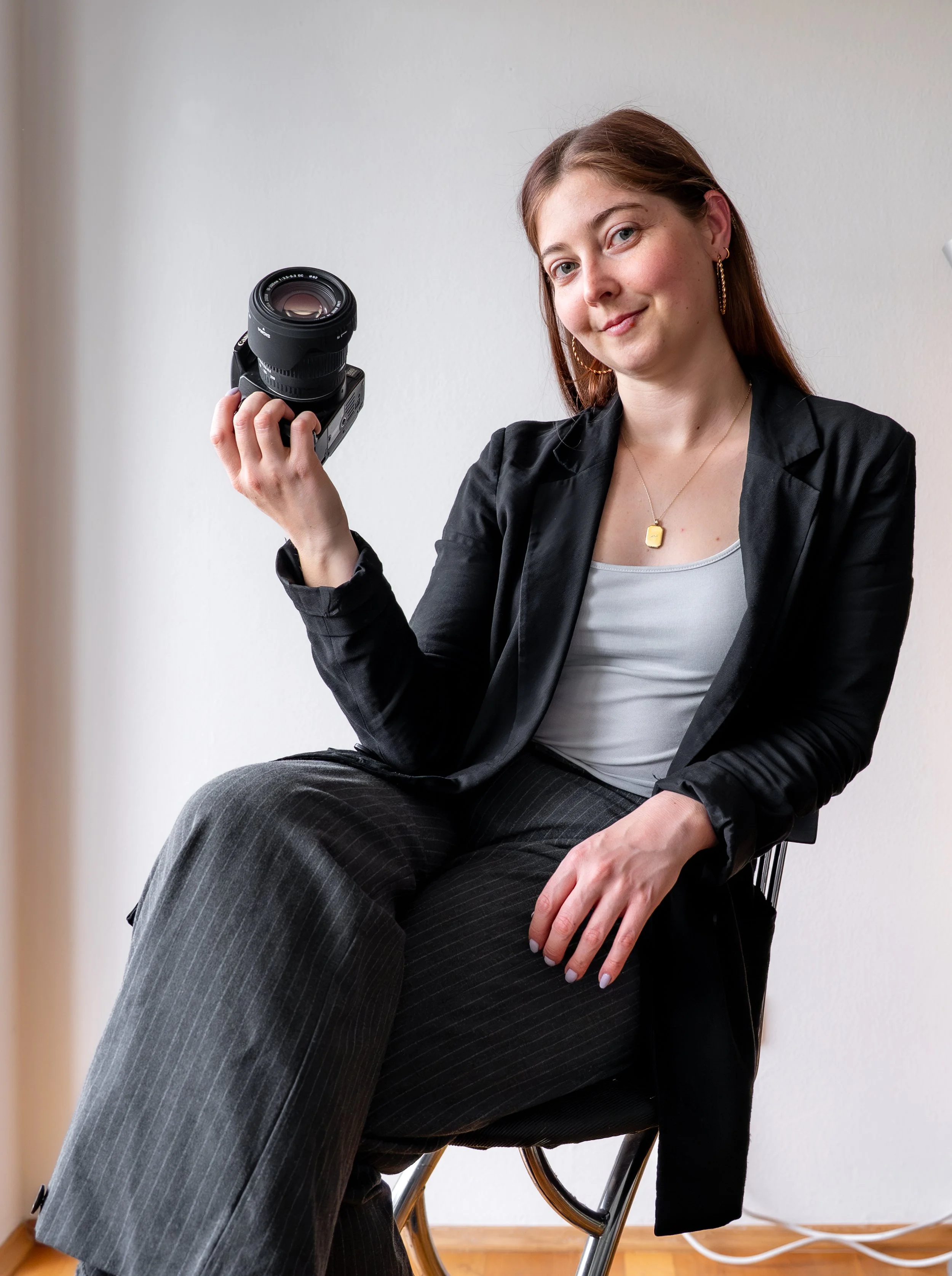Junge Frau sitzt auf einem Stuhl, trägt schwarze Jacke und graue Hosen, hält eine Kamera in der Hand und lächelt leicht.