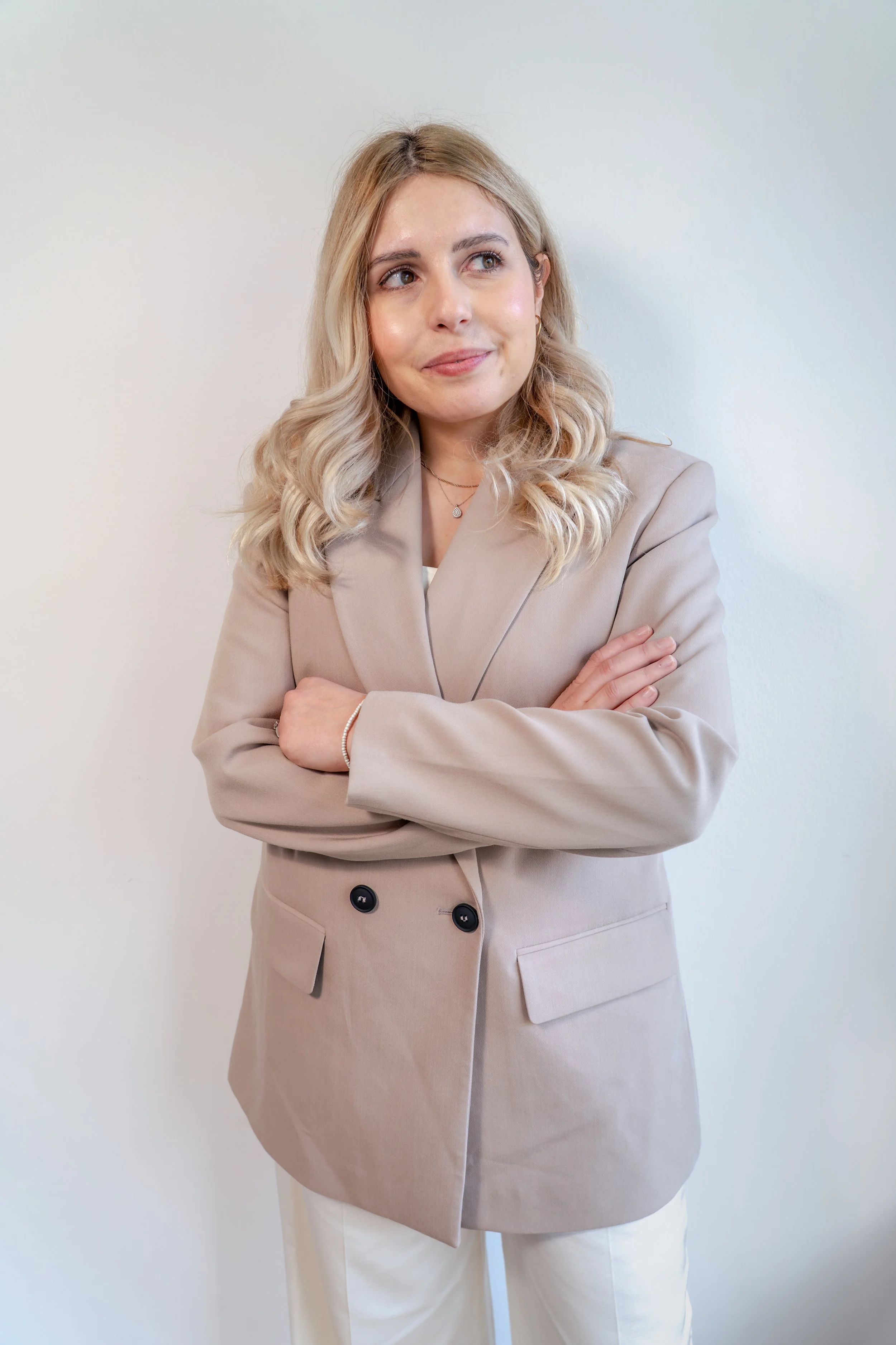 Frau mit blondem, lockigem Haar trägt einen beigen Blazer und weiße Hosen, steht vor weißer Wand, blickt nach rechts, die Arme verschränkt.