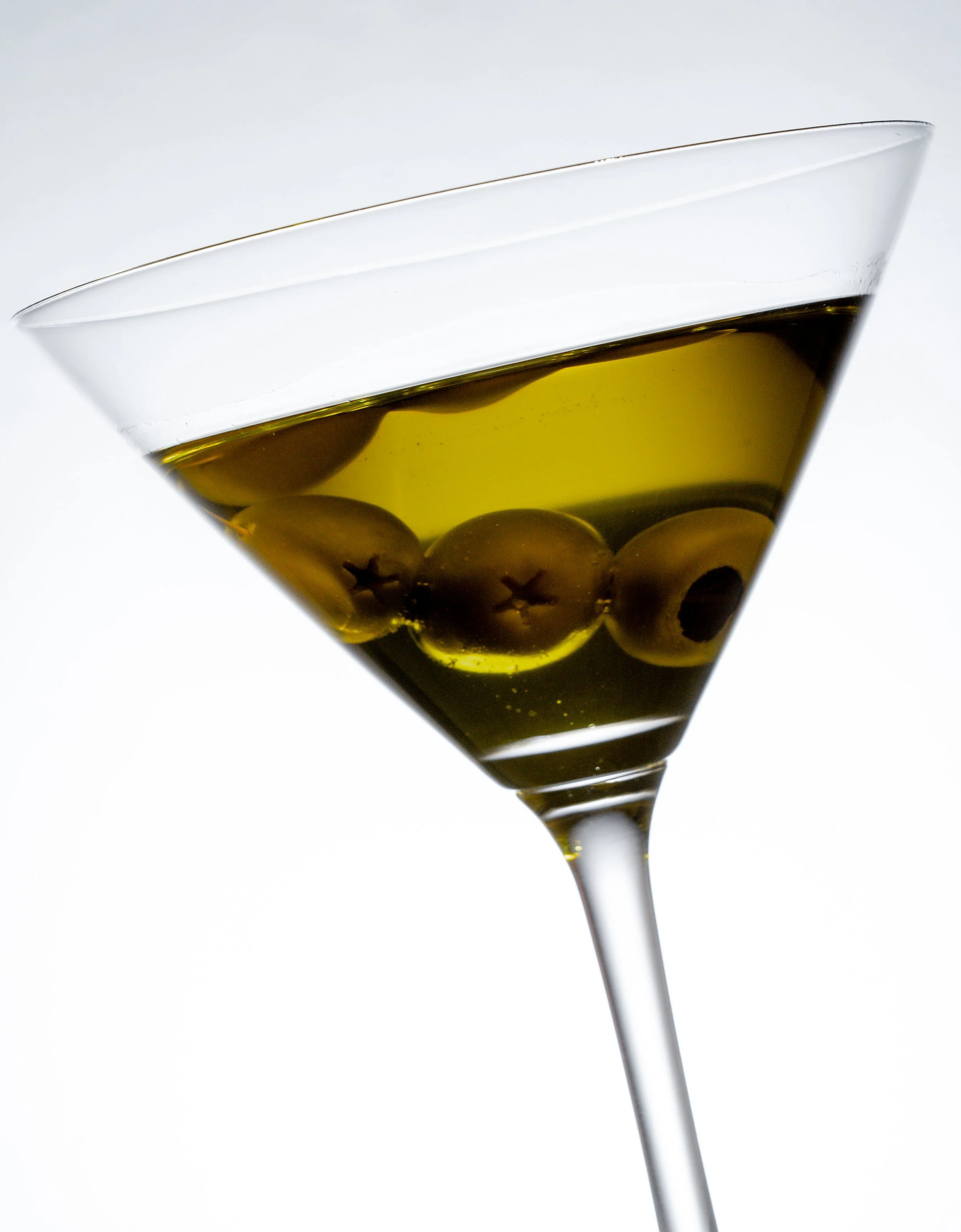Ein Martini-Glas gefüllt mit Olivenöl und grünen Oliven, das vor einem weißen Hintergrund steht.