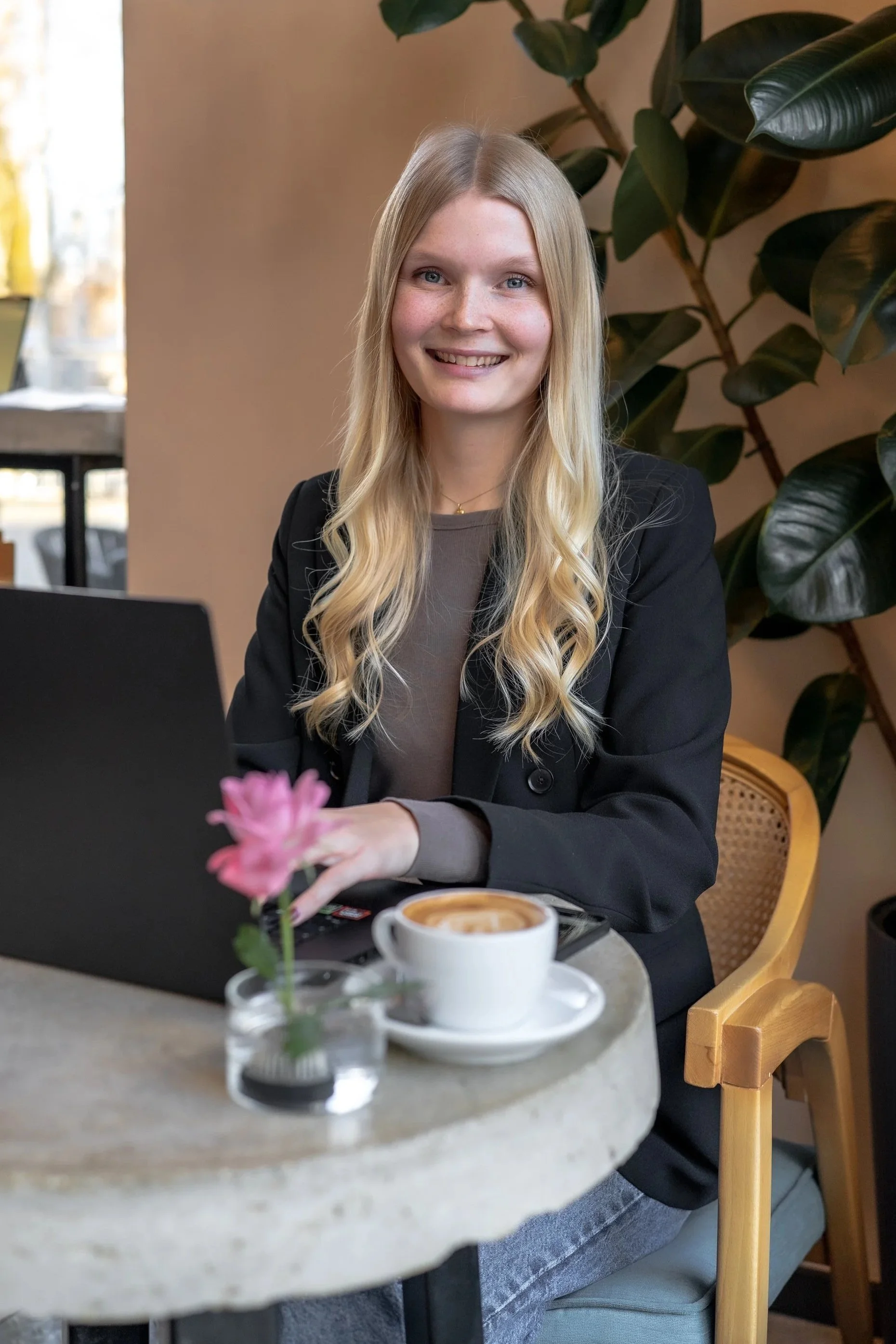 Junge Frau mit blonden, gewellten Haaren sitzt in einem Café vor einem Laptop. Im Vordergrund befindet sich eine Tasse Kaffee und eine kleine Vase mit einer pinken Blume. Im Hintergrund sind große, grüne Pflanzen zu sehen.