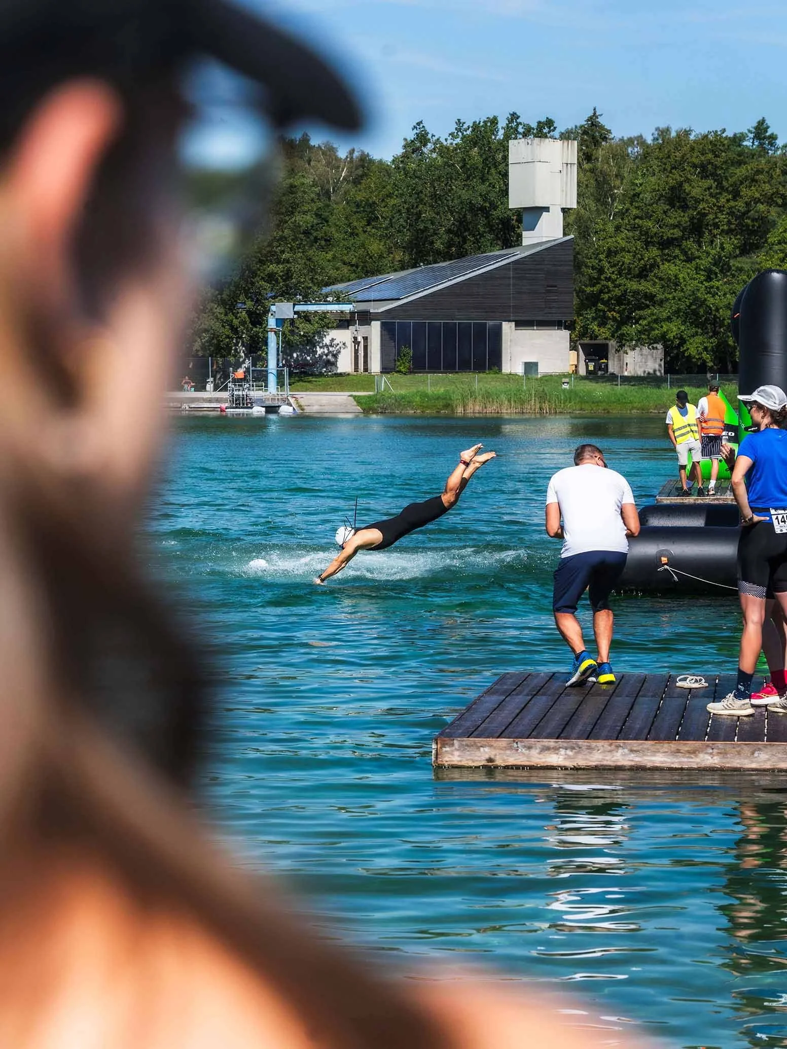 Menschen beim Wassersport am Wasser, inklusive einem Mann im Triathlonanzug, der von einem Steg aus ins Wasser springen, während andere am Pier stehen, im Hintergrund ein Gebäude und Bäume, der Himmel ist klar.