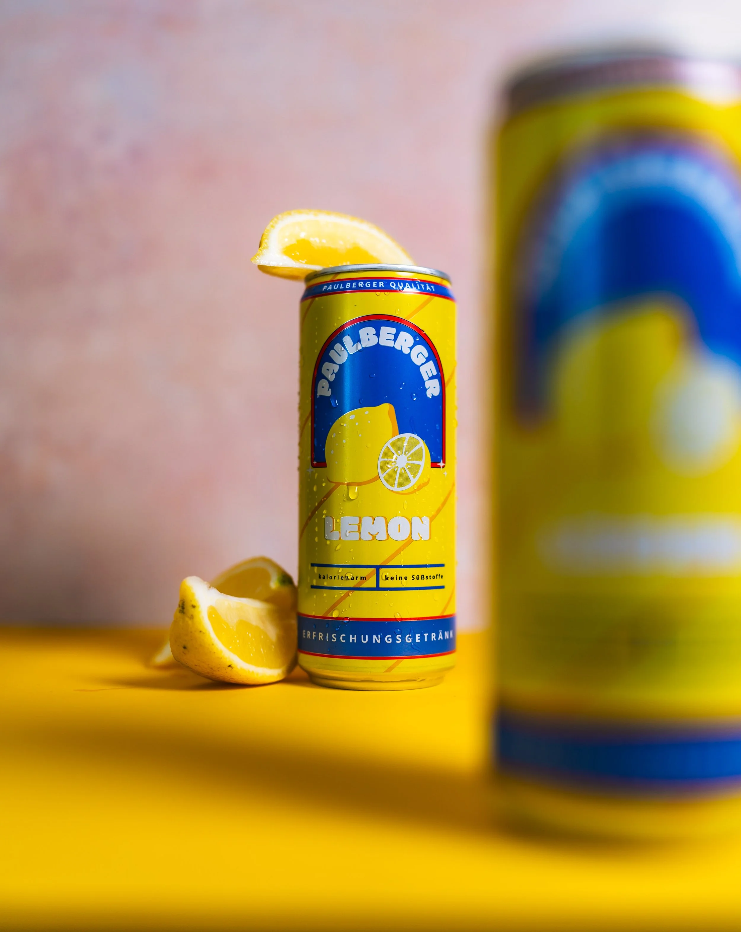Eine gelbe Zitronenlimonade-Dose mit einem Zitronenstück auf der Oberseite, daneben ein halbierte Zitrone. Im Vordergrund ist eine weitere Dosen zu sehen, unscharf.