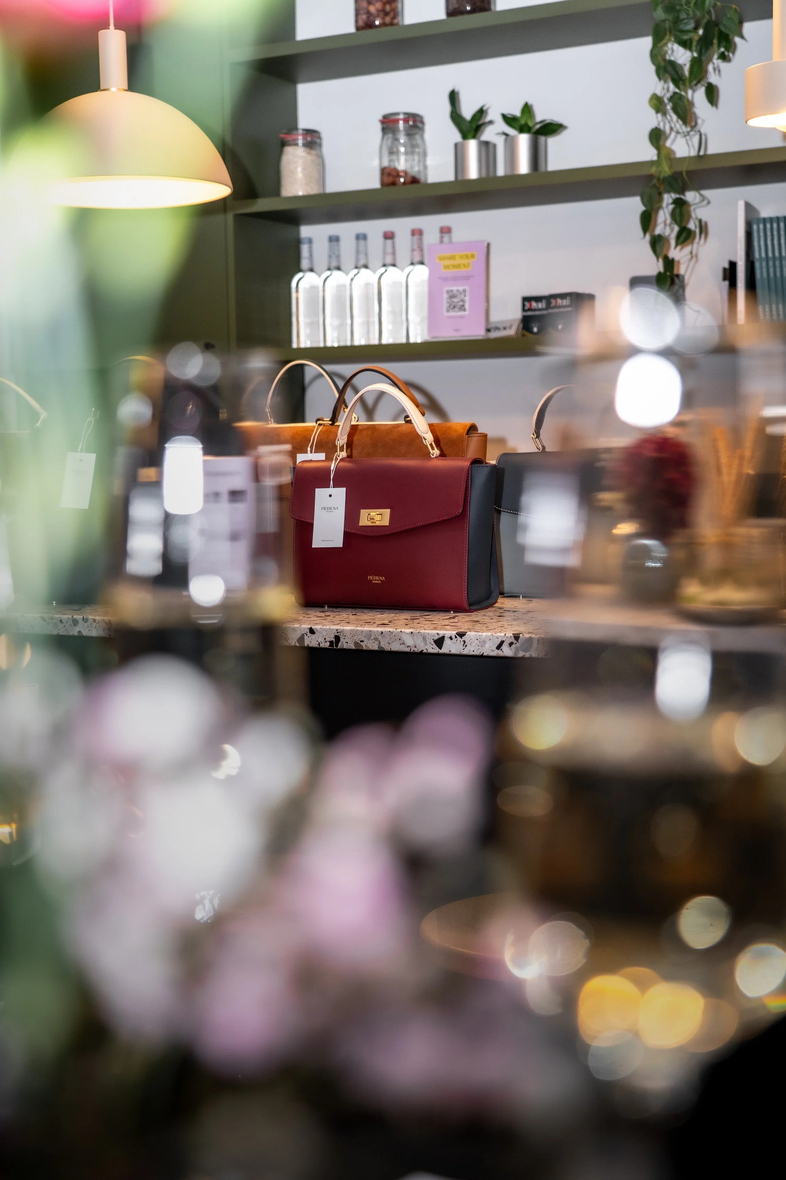 Blumen im Vordergrund, ein Tresen mit Handtaschen und Pflanzen im Hintergrund, Beleuchtung in einem Geschäft.