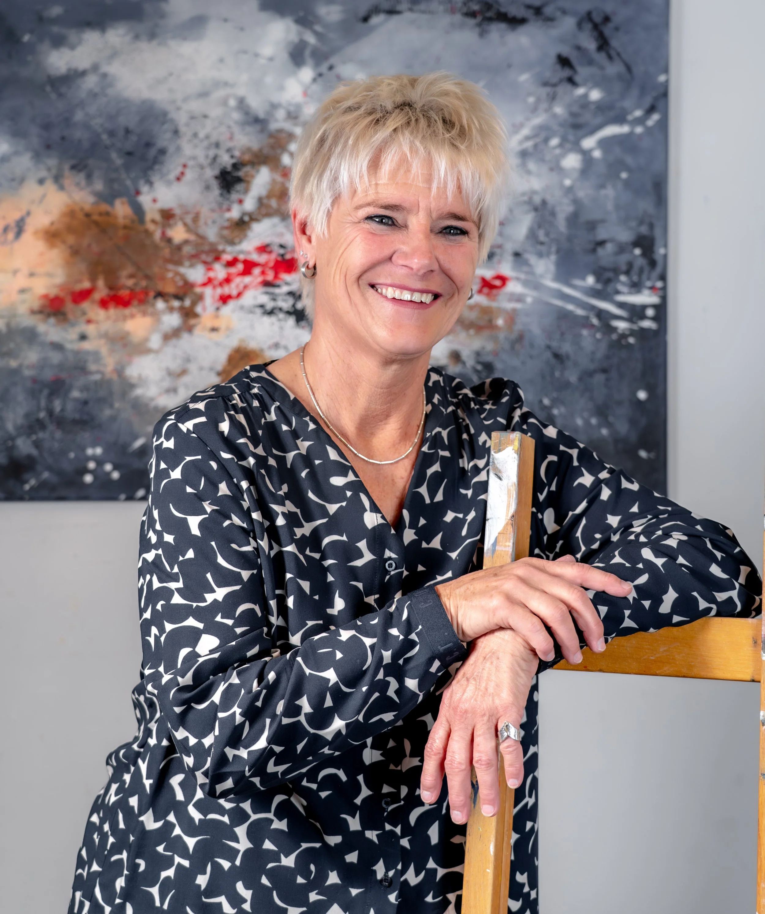 Lächelnde Frau mit kurzer blondem Haar, trägt ein schwarzes Hemd mit weißem Mustern, steht an einer Staffellei mit Pinsel in der Hand vor ihrem gemalten Bild im Hintergrund. Sie ist Künsterlin und das Foto ist in ihrem Studio entstanden.