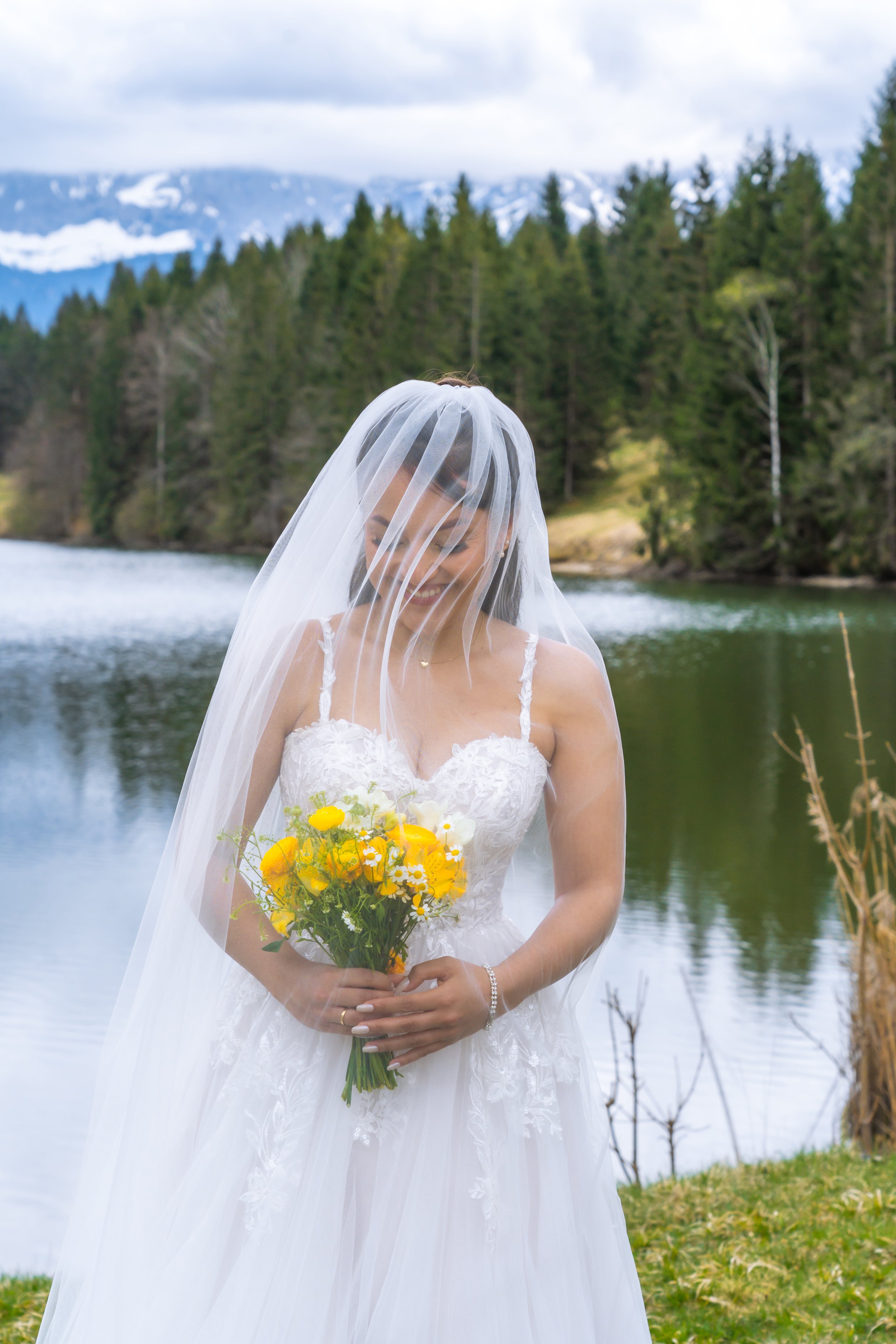 Eine Braut in einem weißen Hochzeitskleid mit Schleier, die einen Strauß gelber und weißer Blumen hält, vor einem See mit Blick auf bewaldete Berge und Wolken am Himmel.