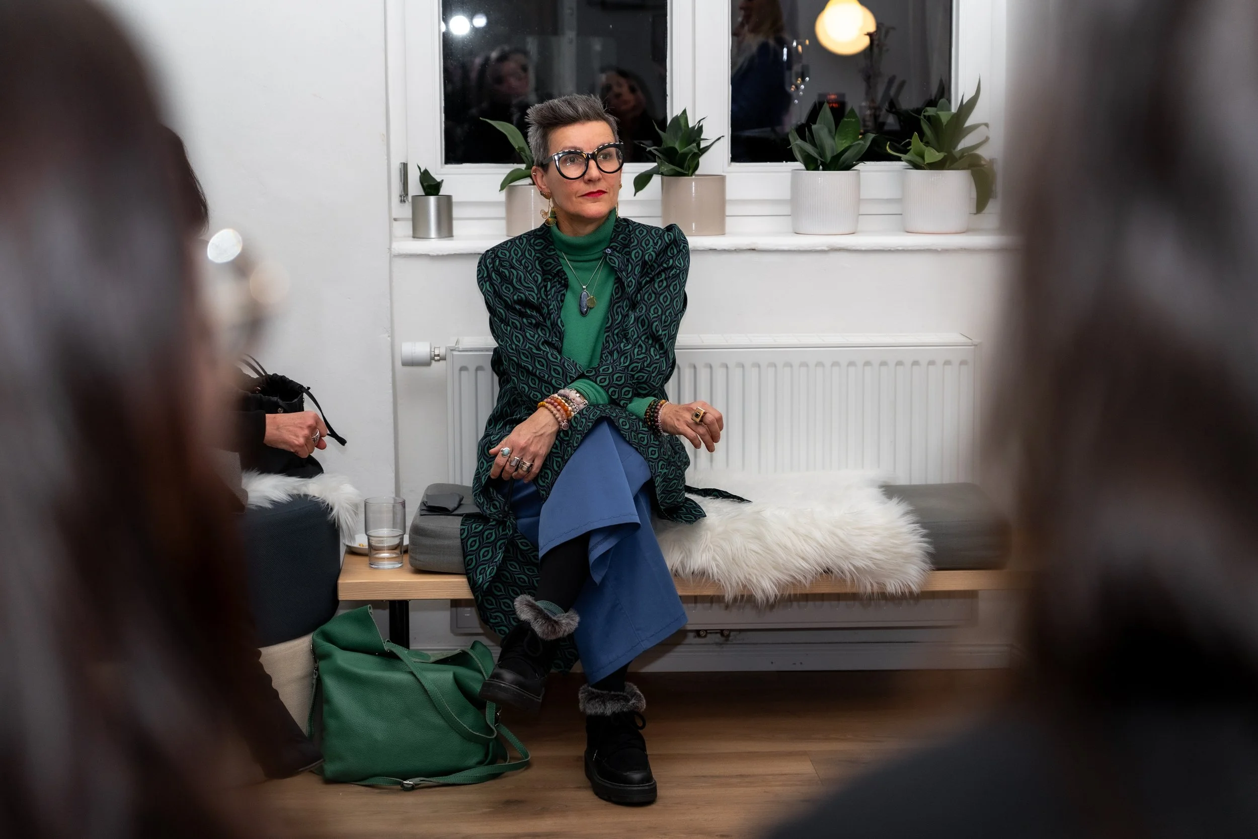 Eine elegant gekleidete Frau mit kürzlichem Grauhaar und großen schwarzen Brillen sitzt an einem Fenster . Sie trägt eine grüne Bluse unter einem schwarzen Musterjacke und mehrere Schmuckstücke. Im Hintergrund sind Pflanzen und ein leerer Heizkörper zu sehen. Die Szene ist warm beleuchtet.