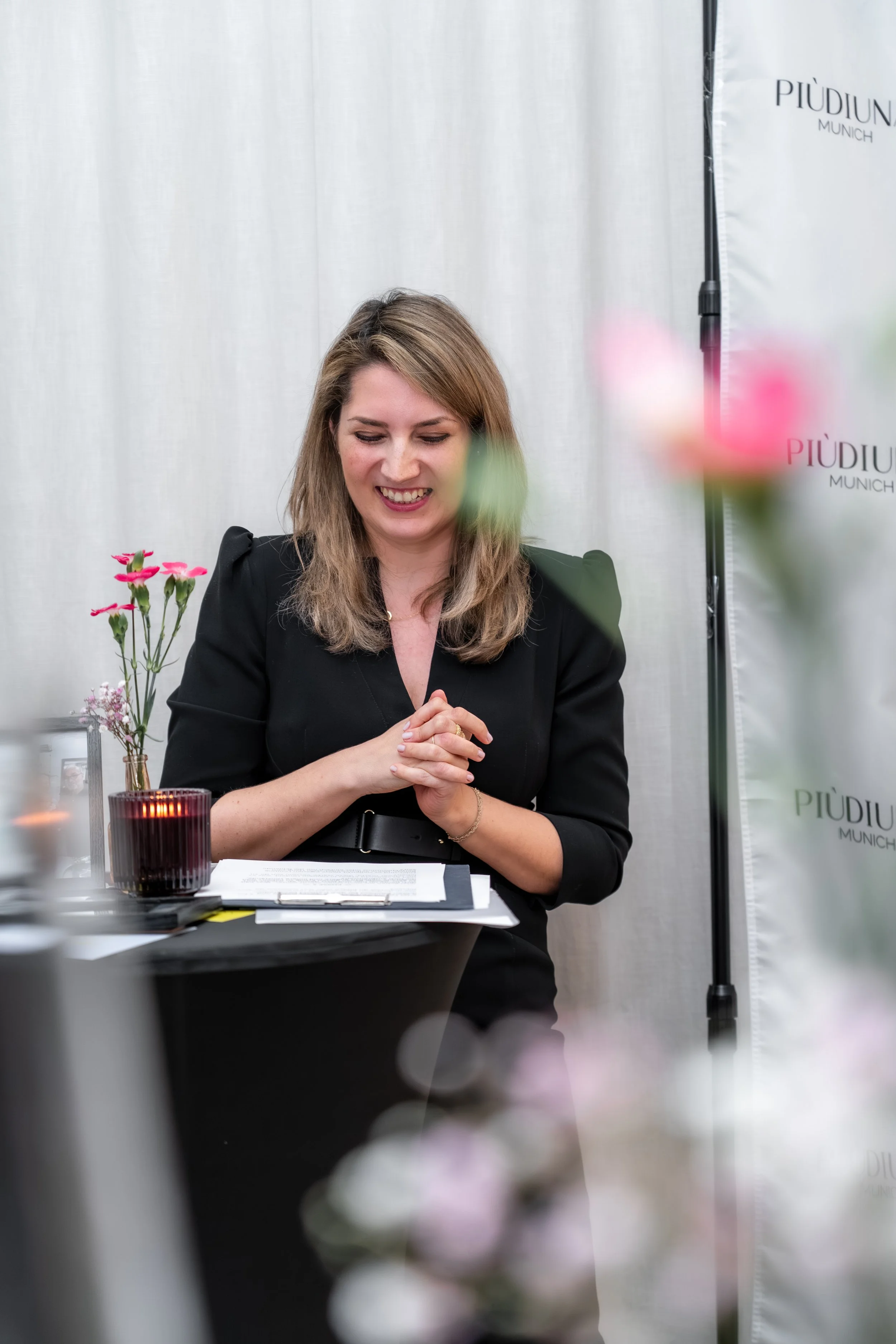Eine lächelnde Frau mit braunen Haaren in einem schwarzen Blazer sitzt an einem Tisch, umgeben von Blumen und Dokumenten, in einem Veranstaltungsraum mit einem weißen Hintergrund und einem PIÙDIUM Logo.