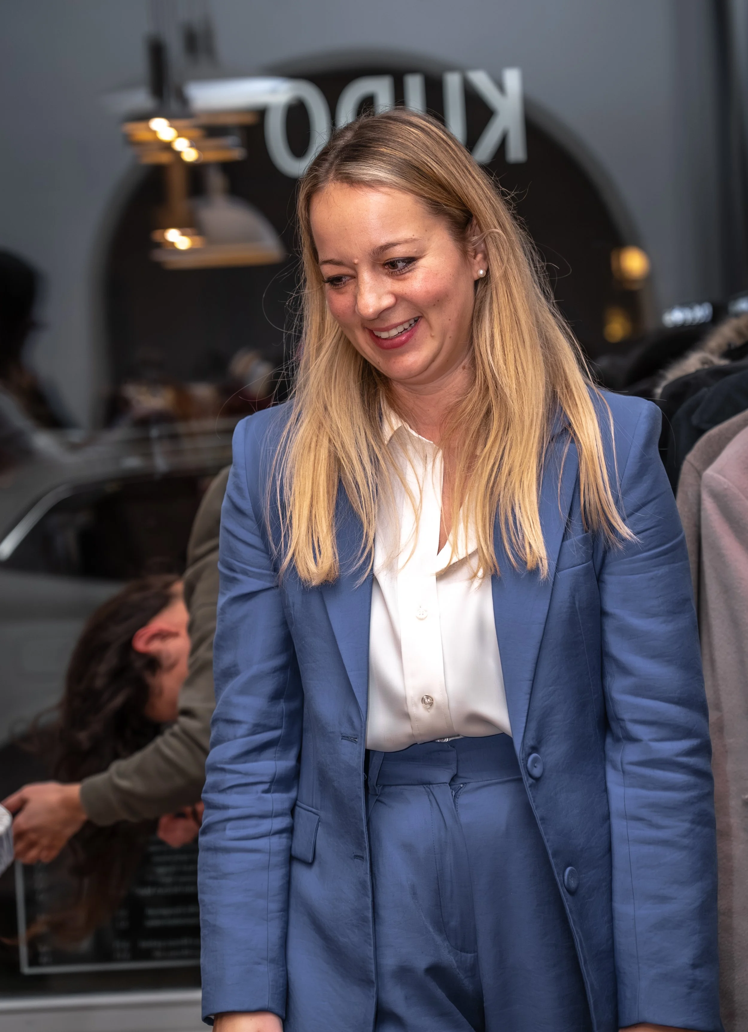 Eine lachende Frau in einem blauen Anzug bei einer informellen Zusammenkunft, im Hintergrund sind weitere Personen und ein Spiegel mit der Aufschrift 'KINO' sichtbar.