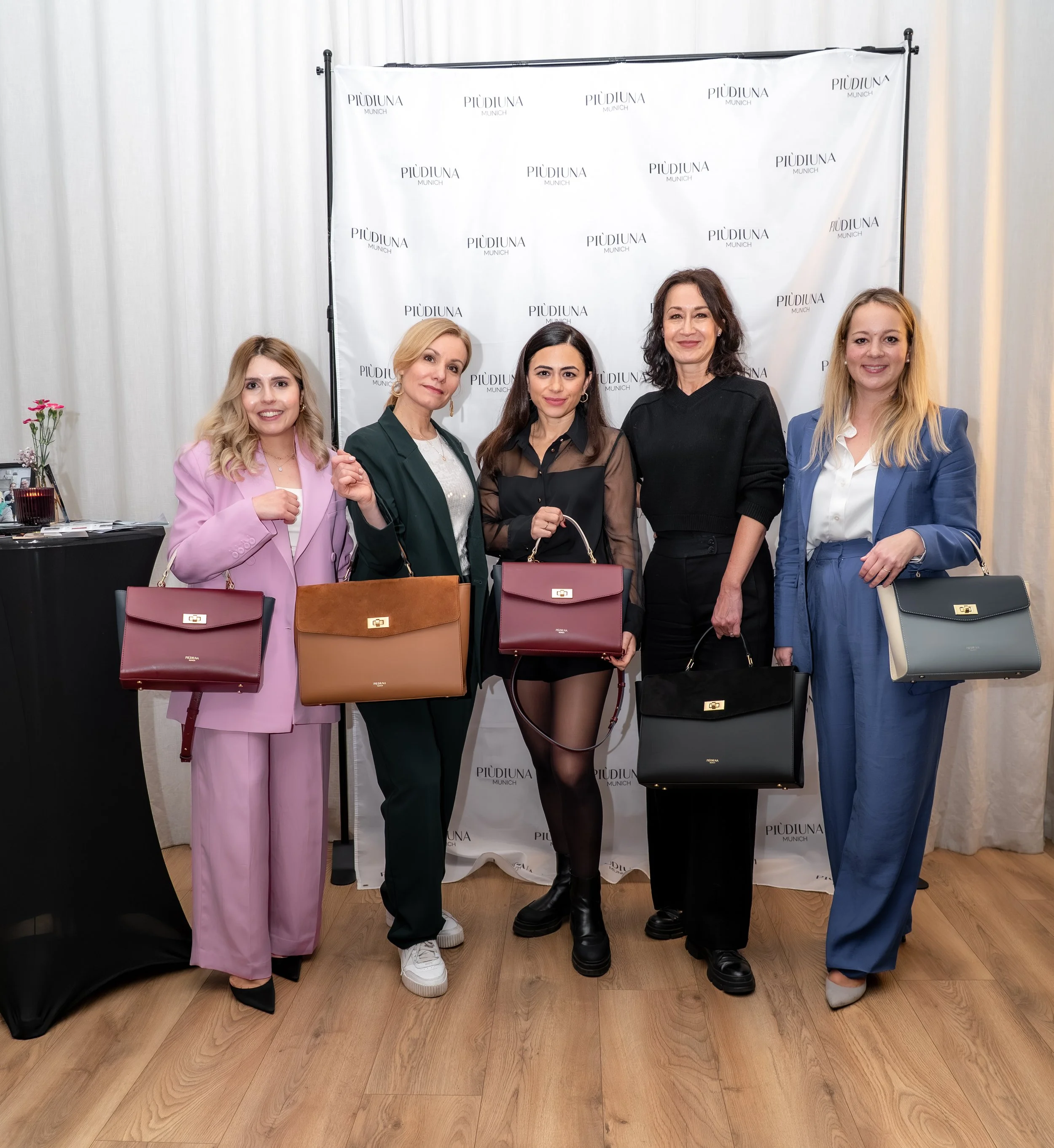 Fünf Frauen stehen nebeneinander vor einem weißen Hintergrund mit dem Schriftzug 'PIÙDIUNA Munich'. Sie posieren für ein Gruppenfoto, jede hält eine Designerhandtasche in verschiedenen Farben. Sie tragen formelle Kleidung in verschiedenen Farben. Die