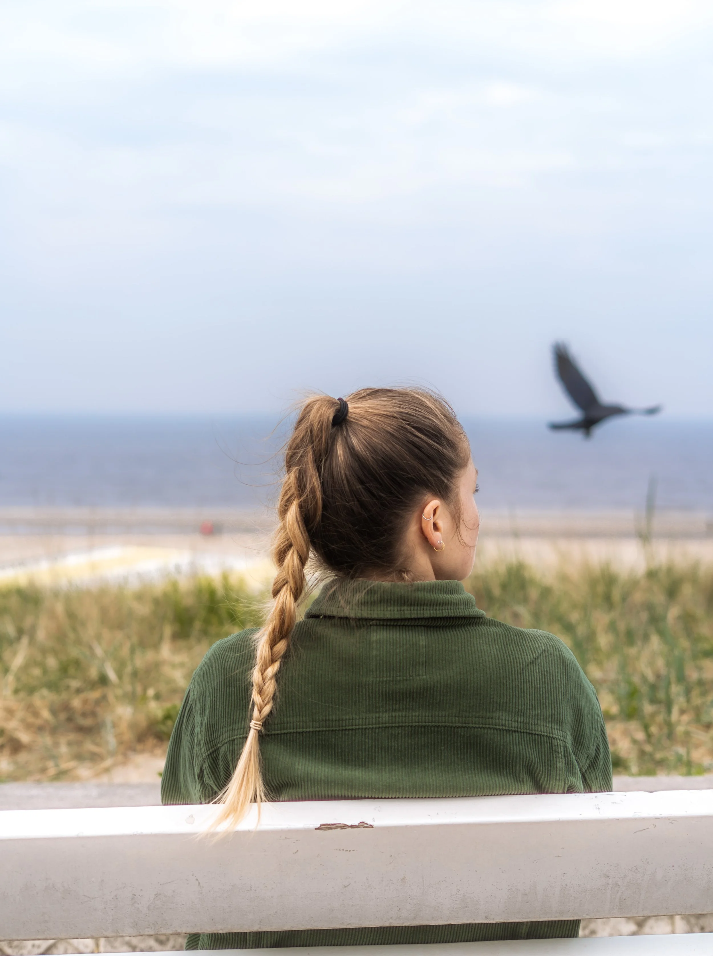 Frau mit geflochtenem Haar sitzt auf einer Bank und blickt auf das Meer, im Hintergrund fliegt ein Vogel.