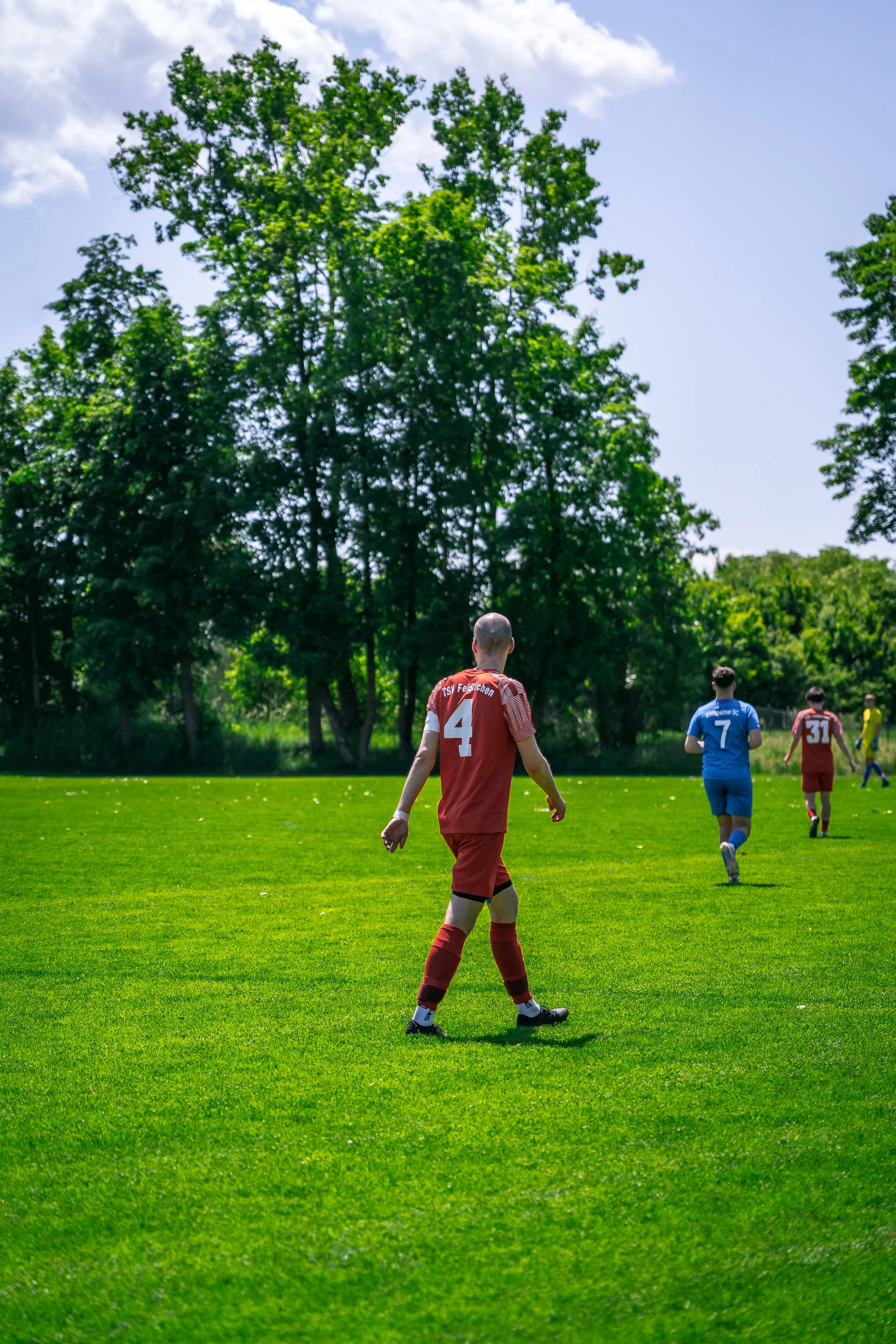Fußballspieler auf grünem Spielfeld an einem sonnigen Tag, mit Bäumen im Hintergrund.