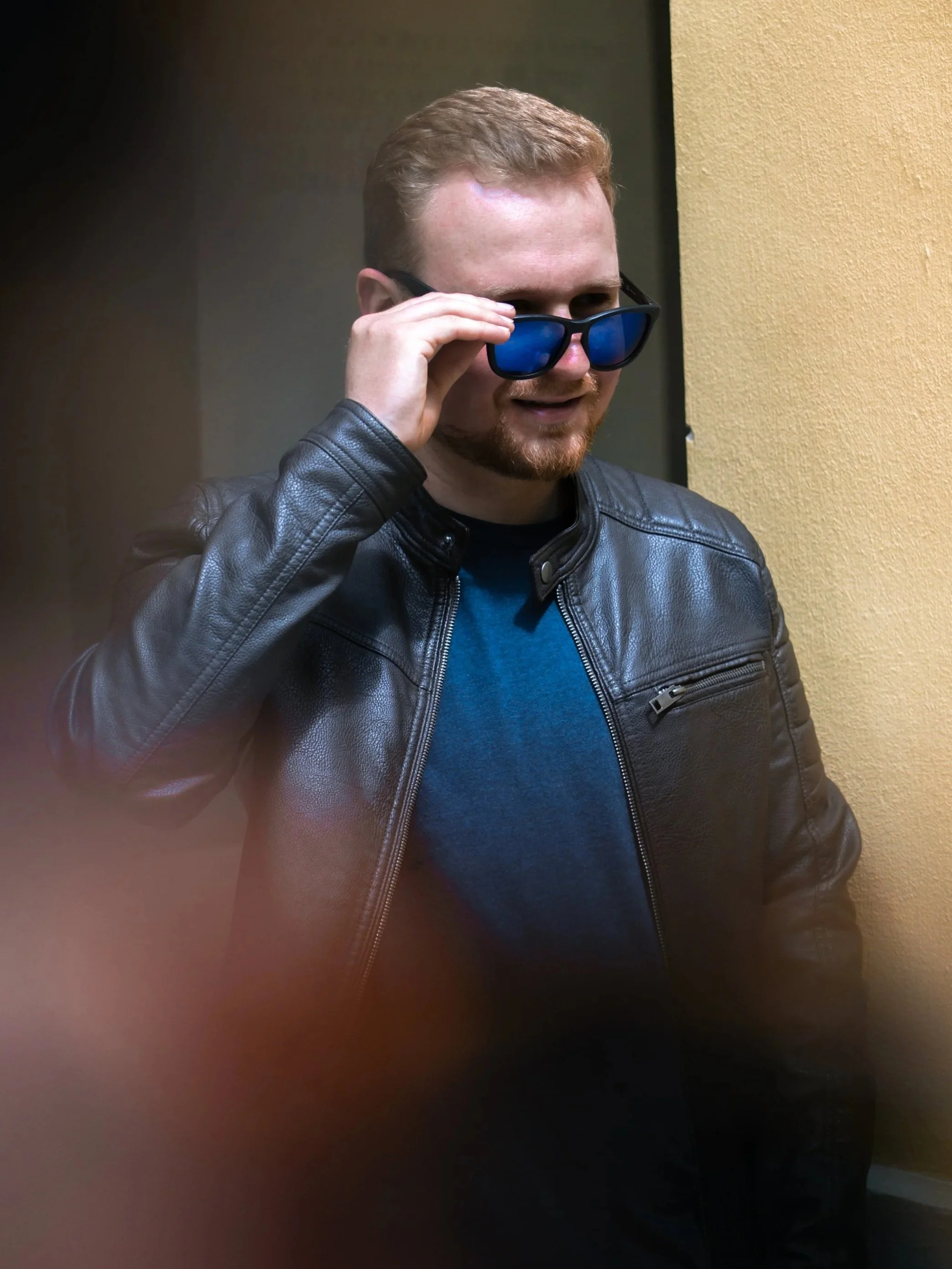 Ein Mann mit rotem Haar, Bräunlichem Bart und dunklen Sonnenbrillen, trägt eine schwarze Lederjacke und steht vor einer gelben Wand.